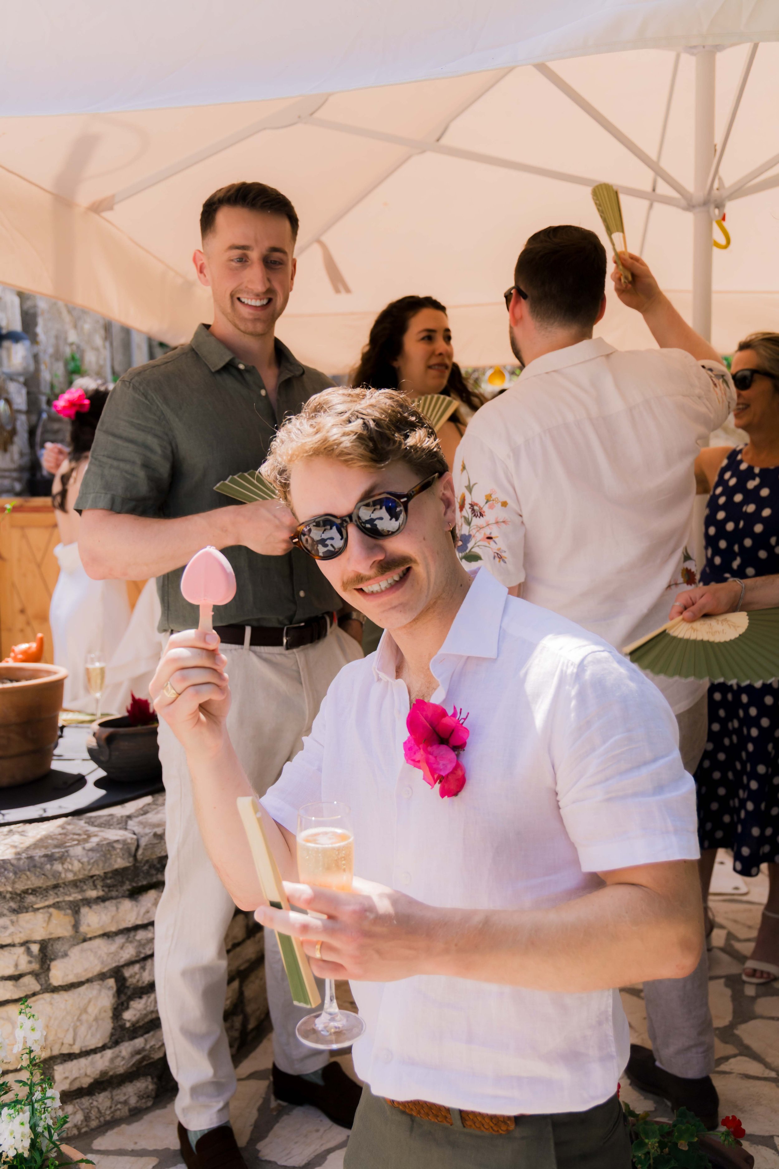 Group of people at a celebration, some holding fans and drinks, with a man in the foreground holding a champagne glass and a pink popsicle, wearing sunglasses and a pink flower boutonniere.
