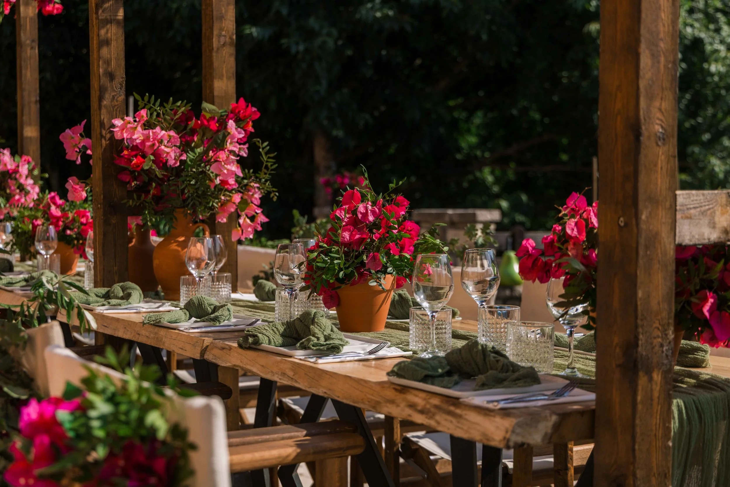 Outdoor dining table decorated with pink flowers in terracotta vases, green napkins, wine glasses, and water glasses, set amid a natural wooden pergola and lush greenery.