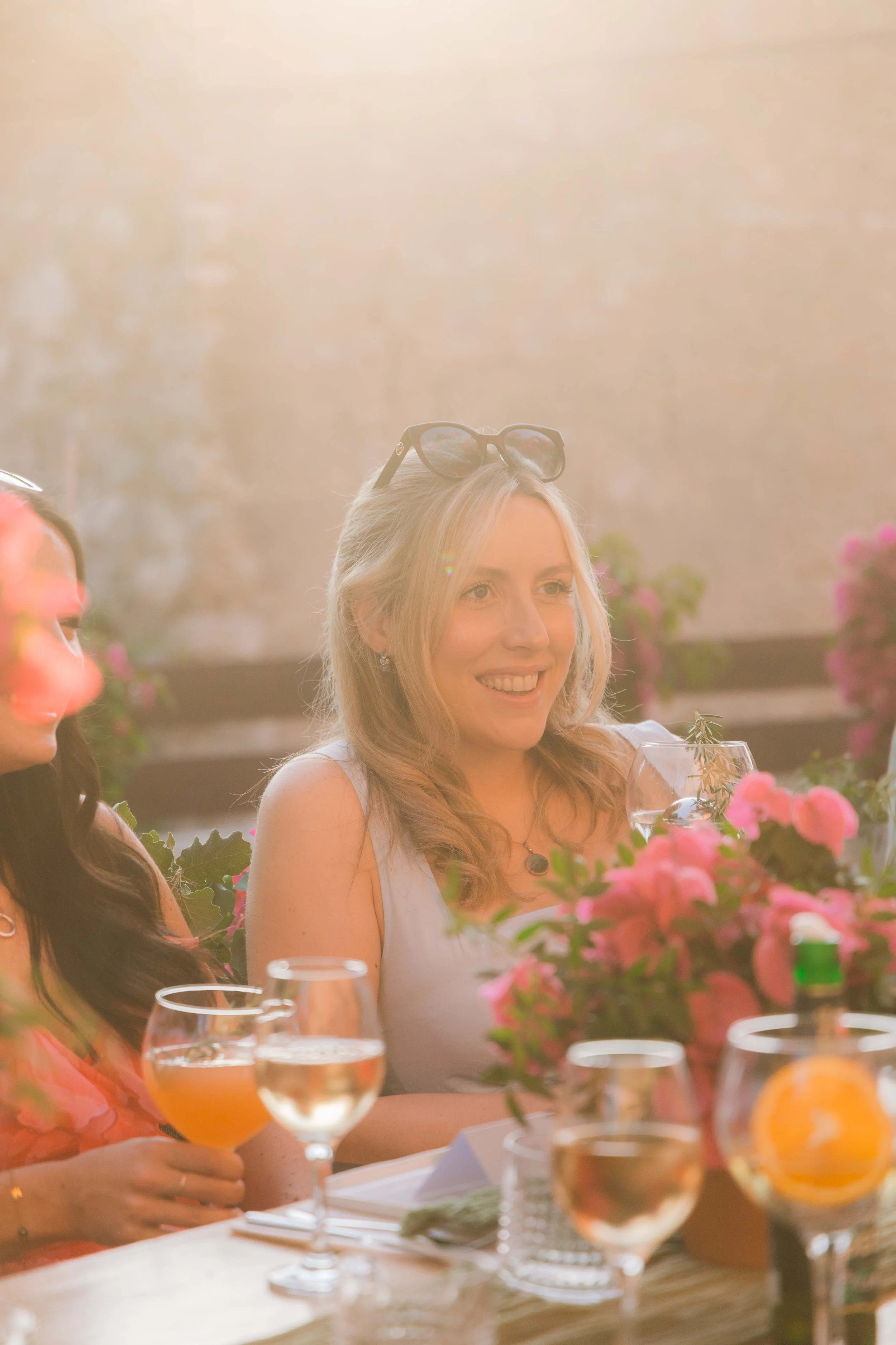 A woman with blonde hair, wearing a light blue top, sitting at a table with wine glasses, flowers, and a glass of water, smiling during an outdoor gathering in the sunlight.