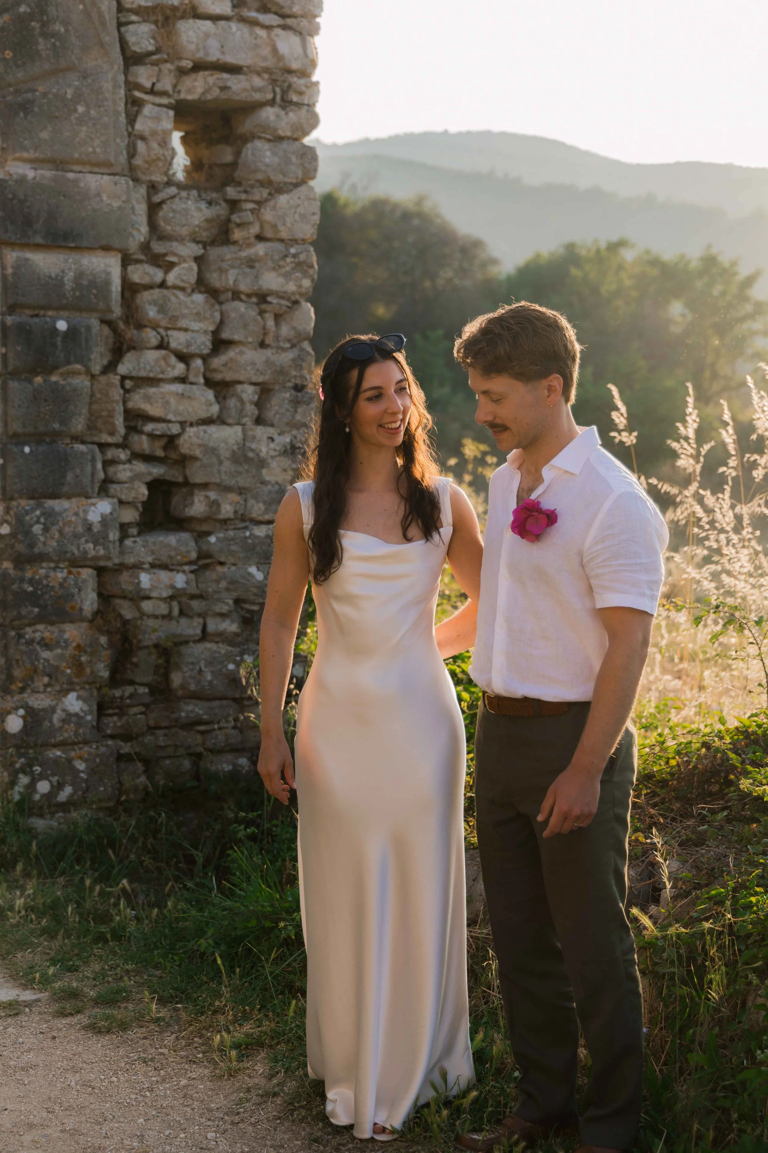 A woman in a white dress and a man in a white shirt with a pink flower on his chest, standing outdoors near a stone structure during sunset.