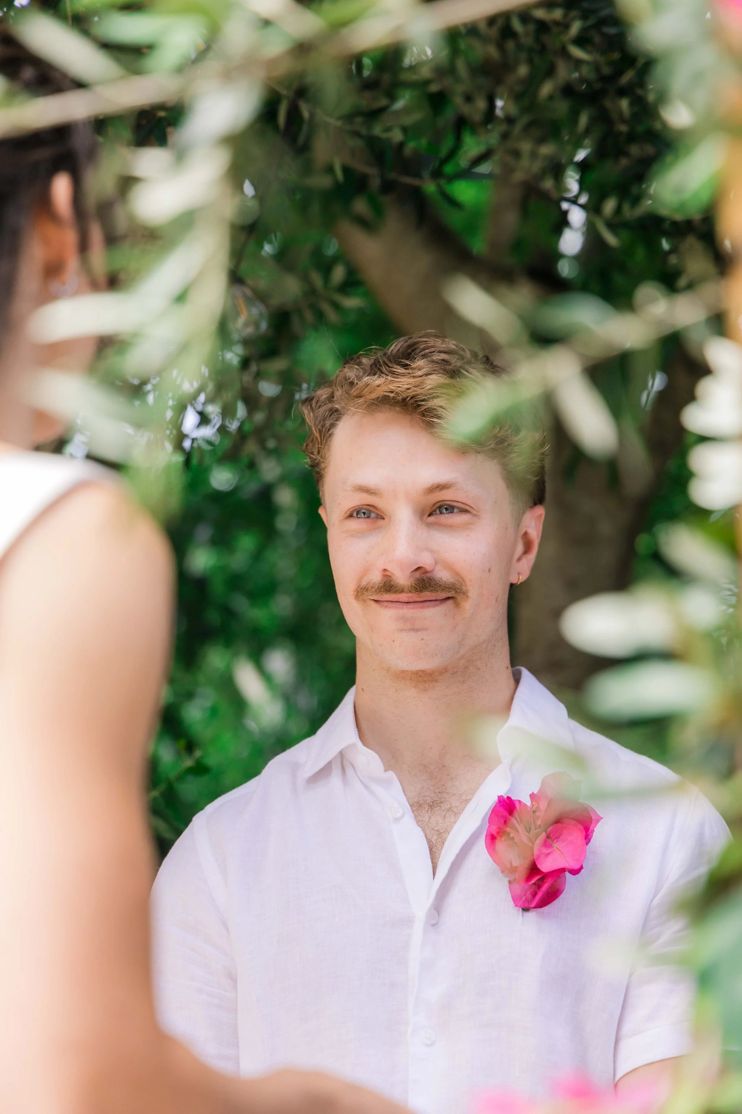 Groom in white shirt with pink boutonniere standing outdoors during wedding, smiling at bride.