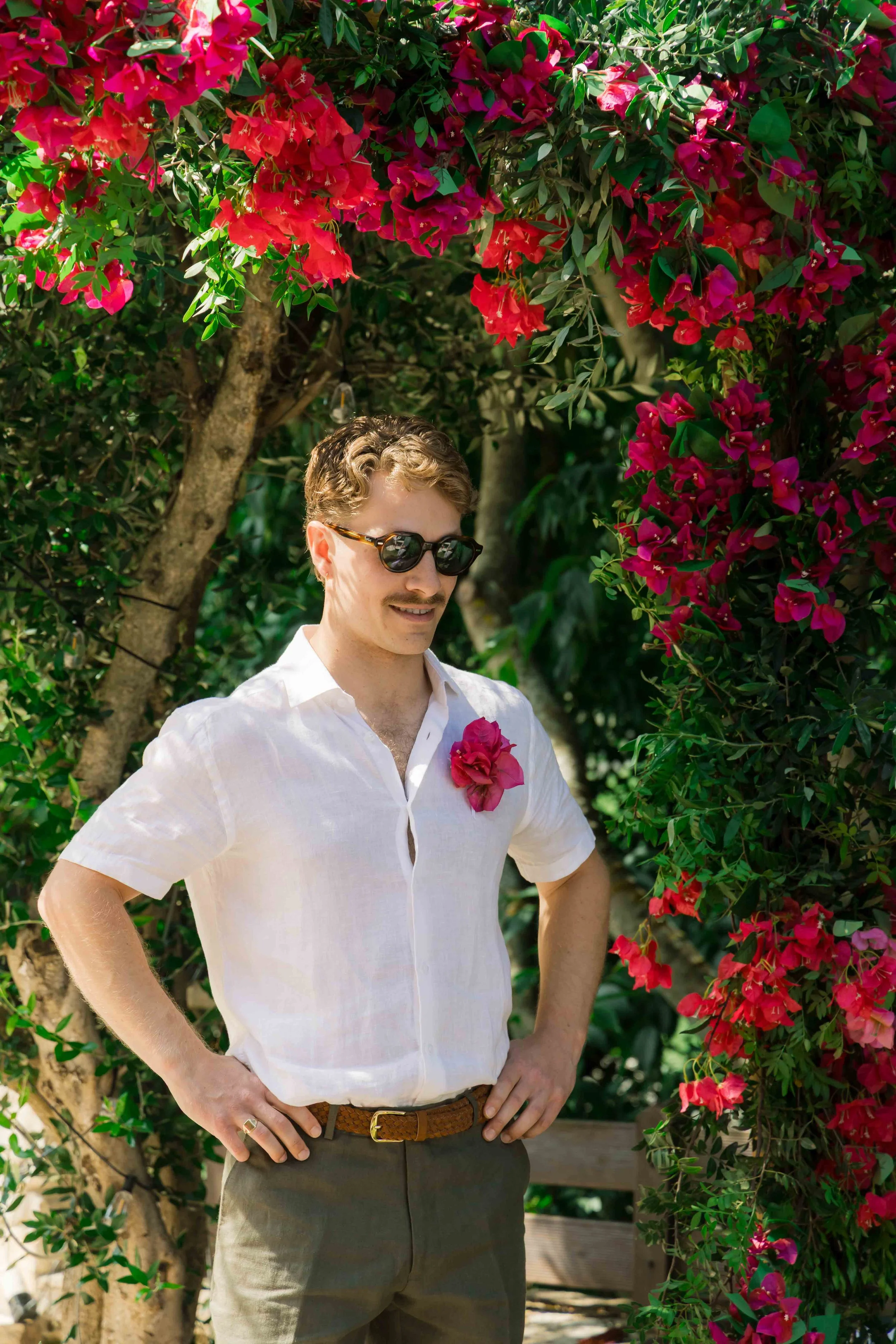 A man in sunglasses wearing a white shirt and brown belt stands outdoors with hands on his hips, surrounded by pink and red flowering bushes.