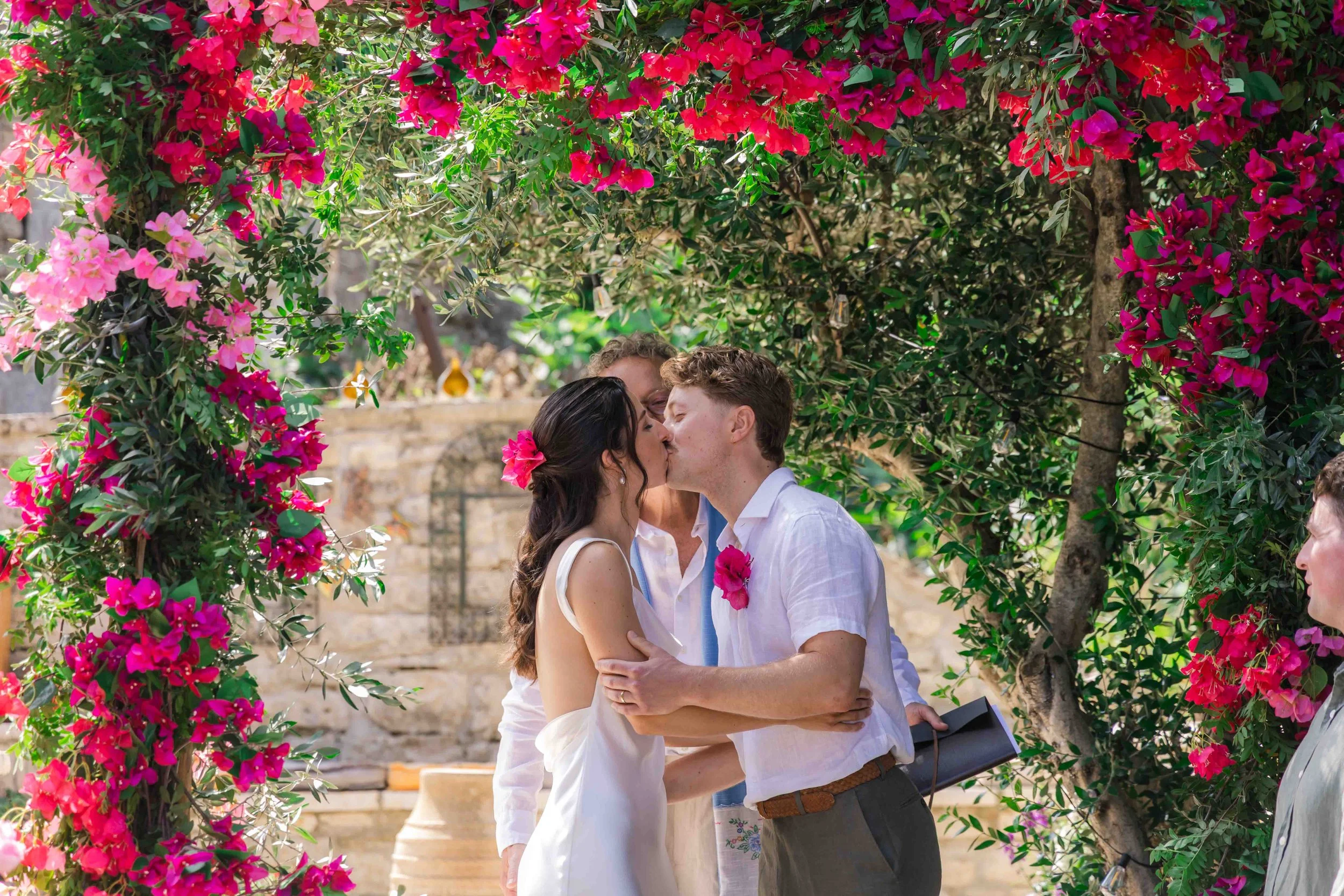 A couple is kissing during their wedding ceremony under a floral arch of pink and red flowers, with an officiant and guests in the background.