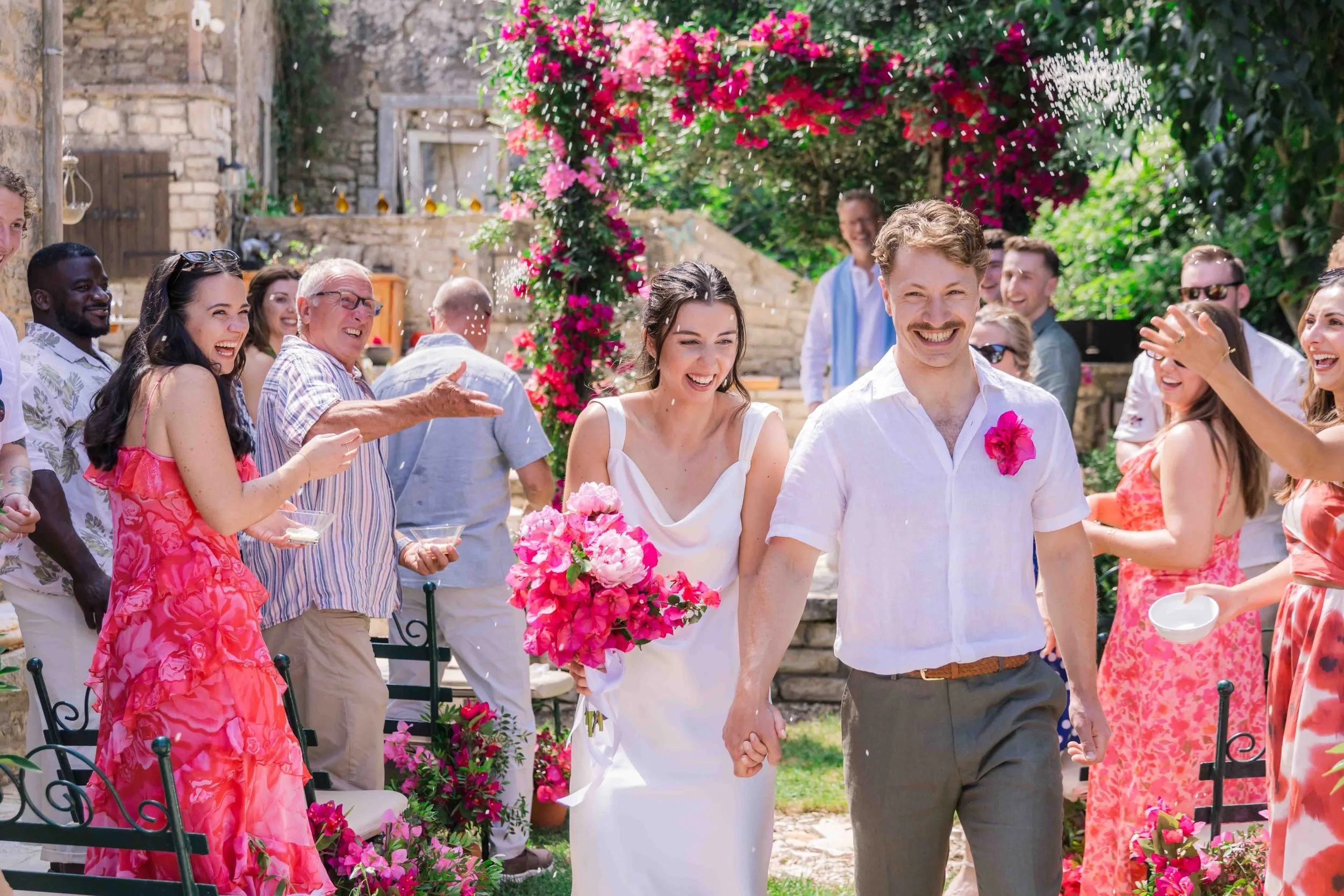 A wedding celebration outdoors with the bride and groom holding hands and smiling, surrounded by friends and family. The bride is holding a bouquet of pink flowers, and everyone appears joyful. There is a floral arch in the background and vibrant gre