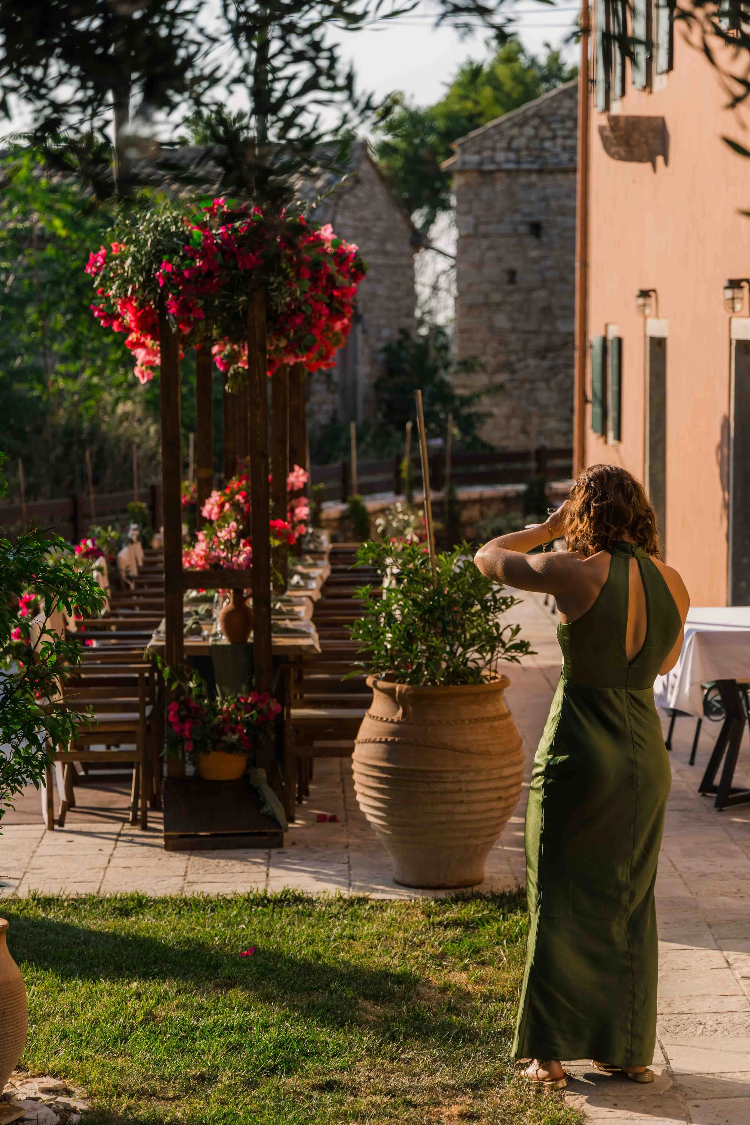 A woman with curly hair wearing a green dress on an outdoor patio surrounded by potted plants and pink flowers, with a building and stone structures in the background.