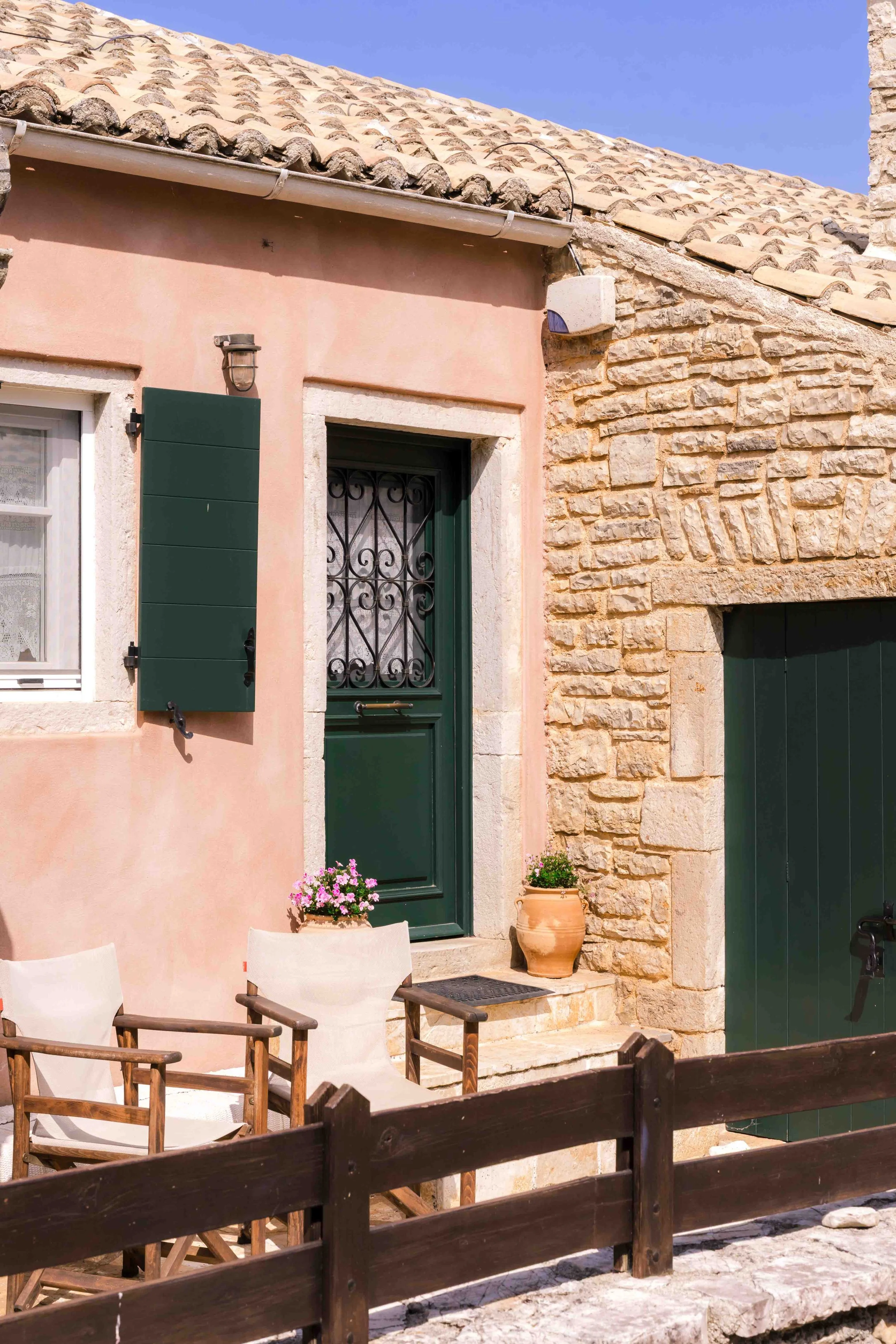 A European-style building with a pink stucco wall and a stone wall, green shutters and door, small outdoor patio with wooden chairs and flowering plants in terracotta pots, wooden fence in the foreground, and clear blue sky.