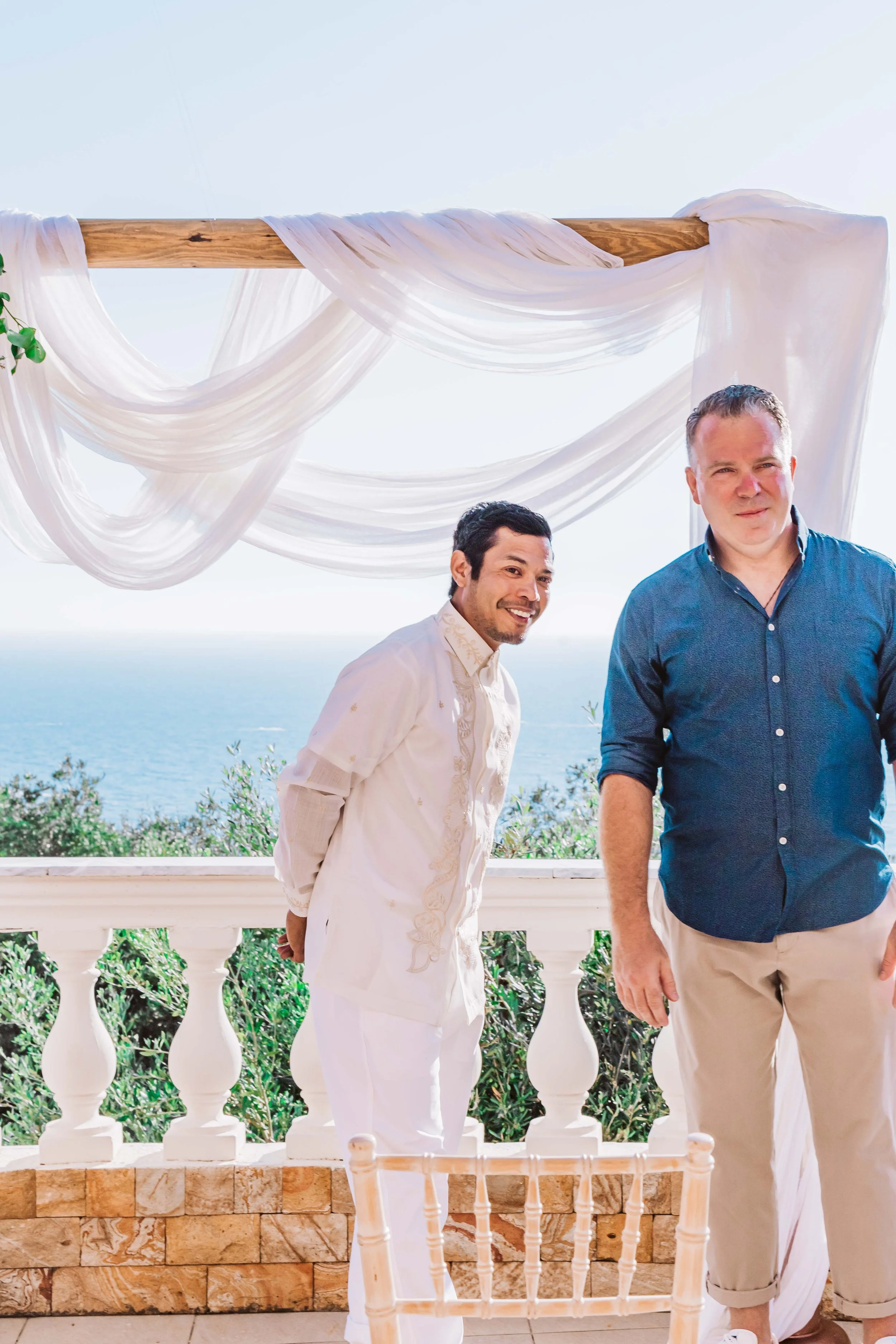 Two men standing on a balcony decorated with white drapes and overlooking the ocean.
