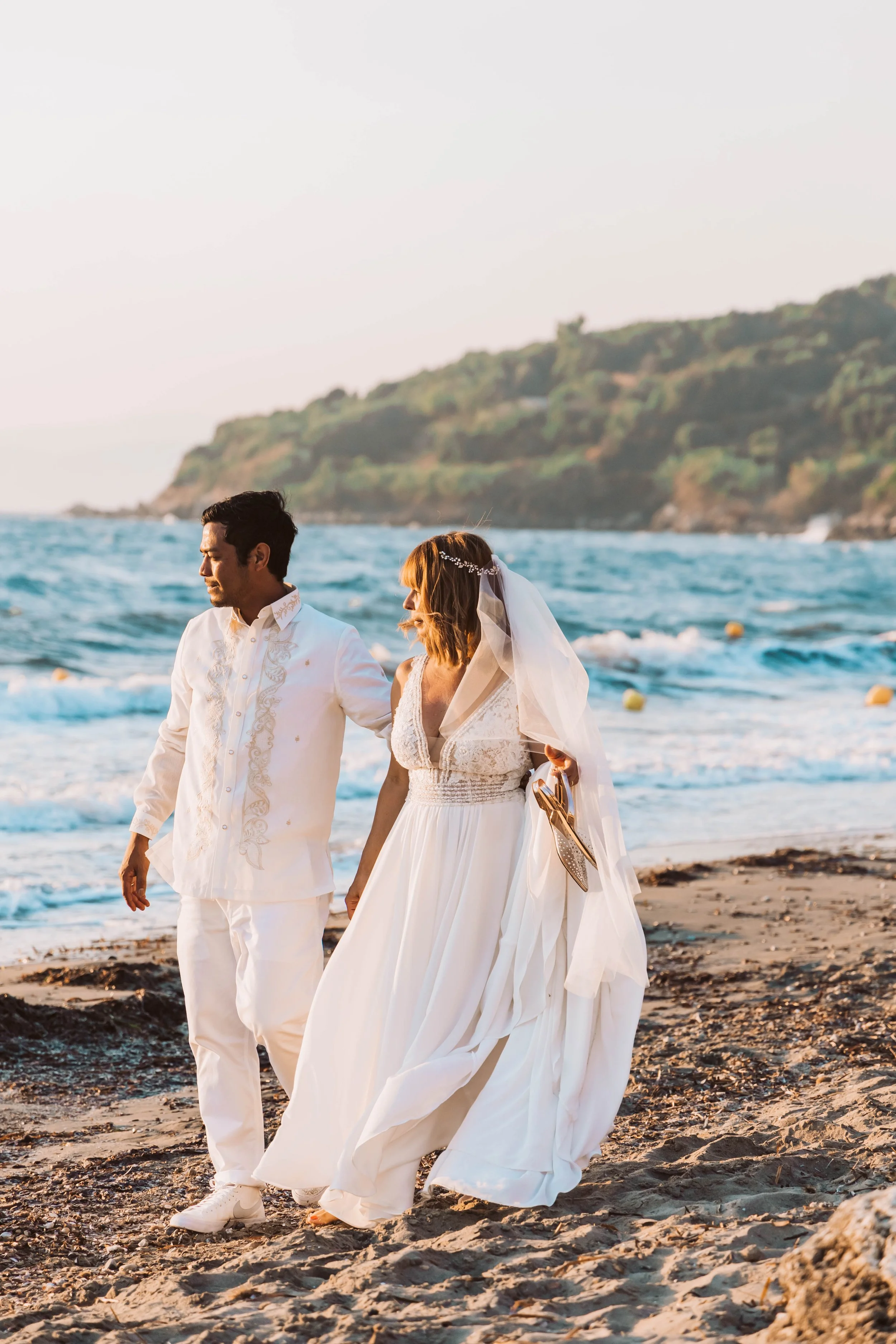 A bride and groom walking on a rocky beach during sunset, with the bride holding her shoes and wearing a white wedding dress and veil, and the groom in a white embroidered suit, with ocean and green hills in the background.