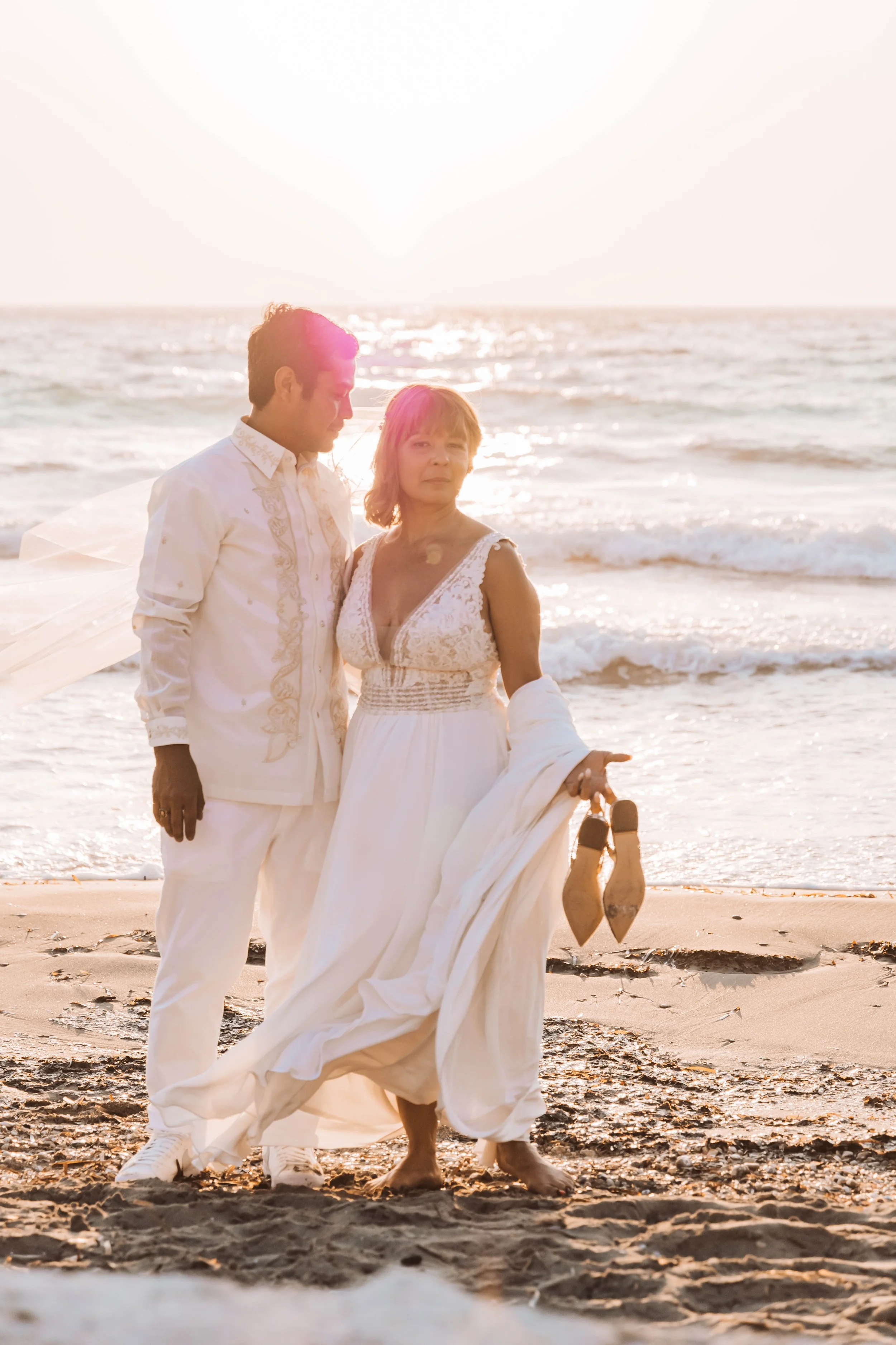 A couple standing on the beach during sunset, dressed in wedding attire, with the woman holding her shoes and the man looking at her.