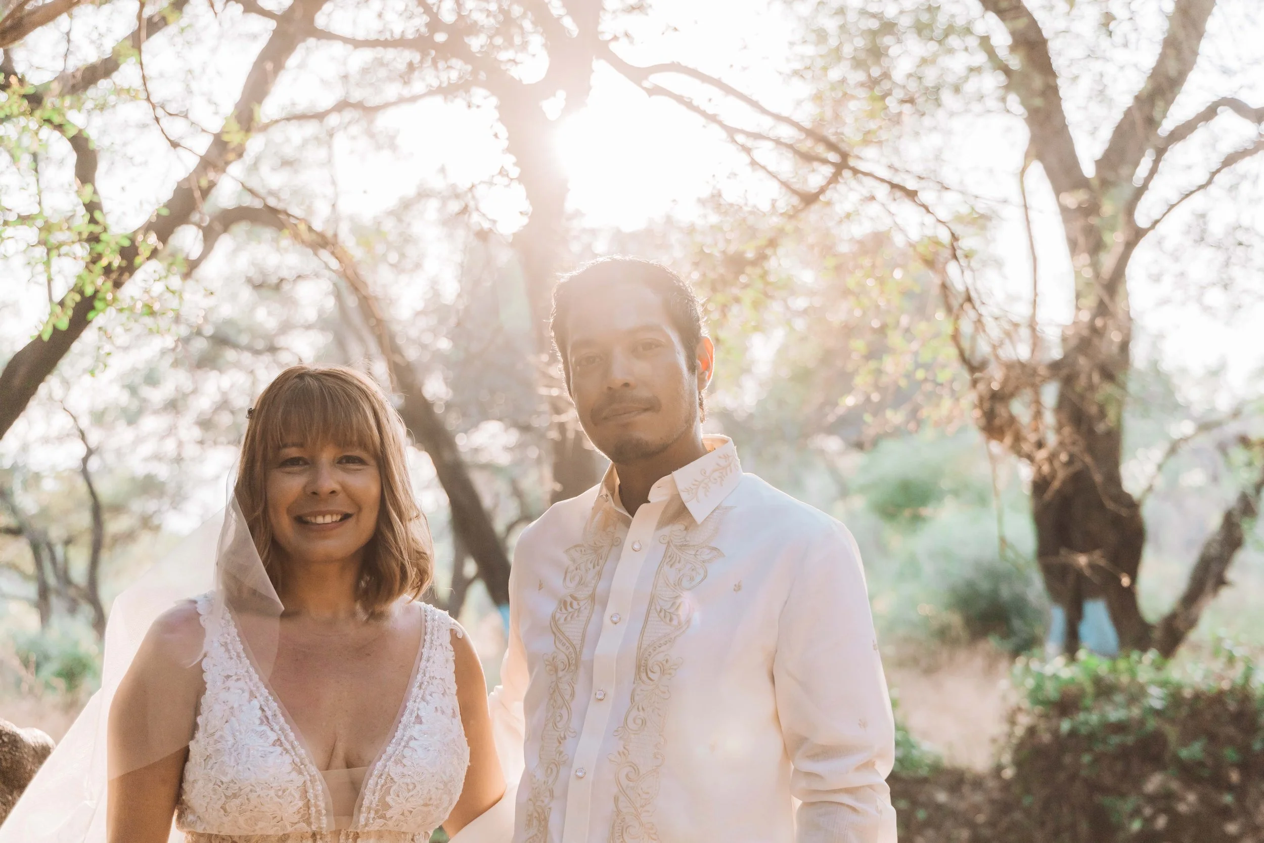 A couple in wedding attire standing outdoors in a wooded area with sunlight streaming through the trees behind them.