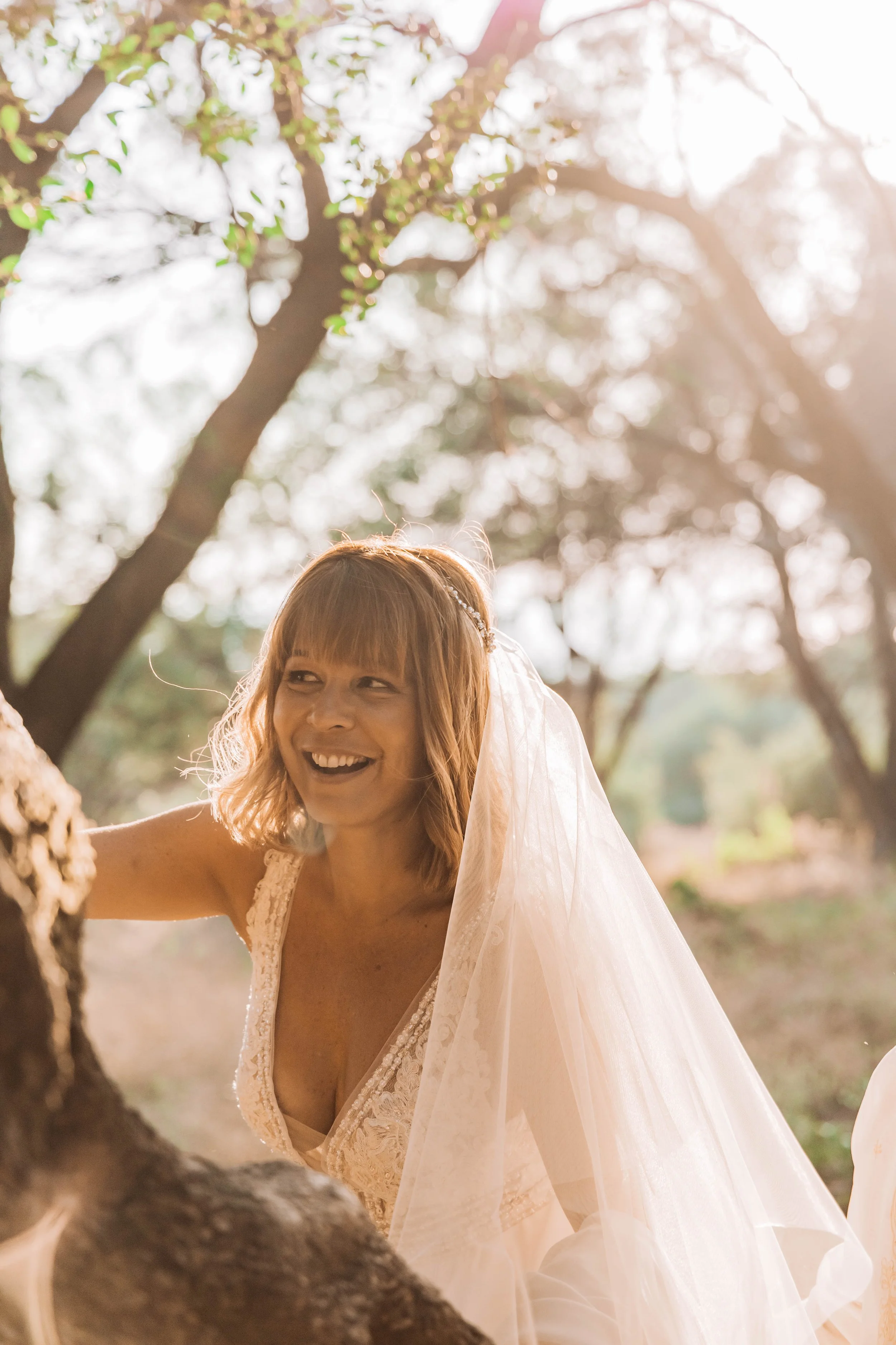 A smiling woman in a wedding dress with a veil outdoors, surrounded by trees with sunlight filtering through the branches.