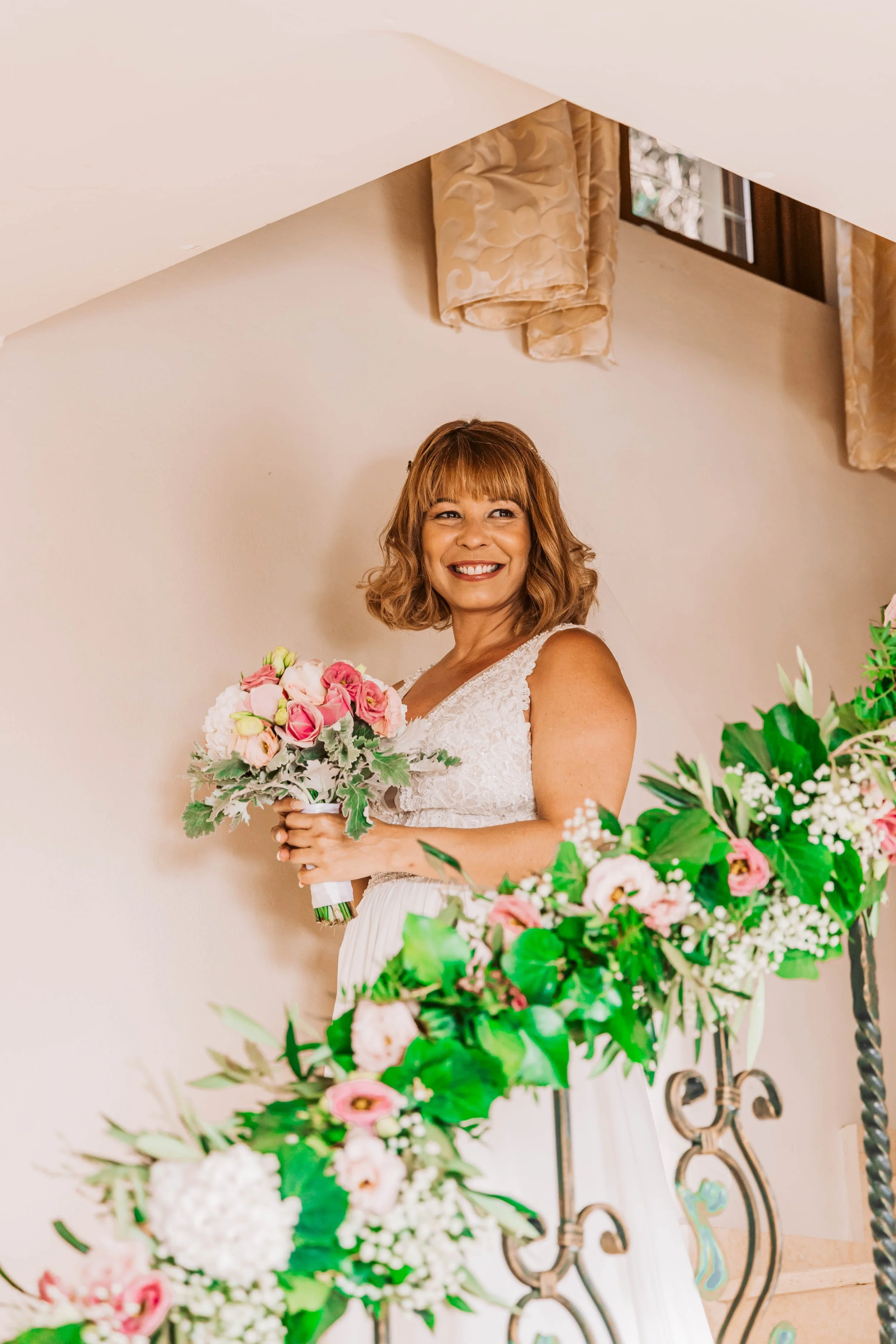 A woman in a wedding dress holding a bouquet of pink and white flowers, smiling indoors near floral decorations and a window with beige curtains.