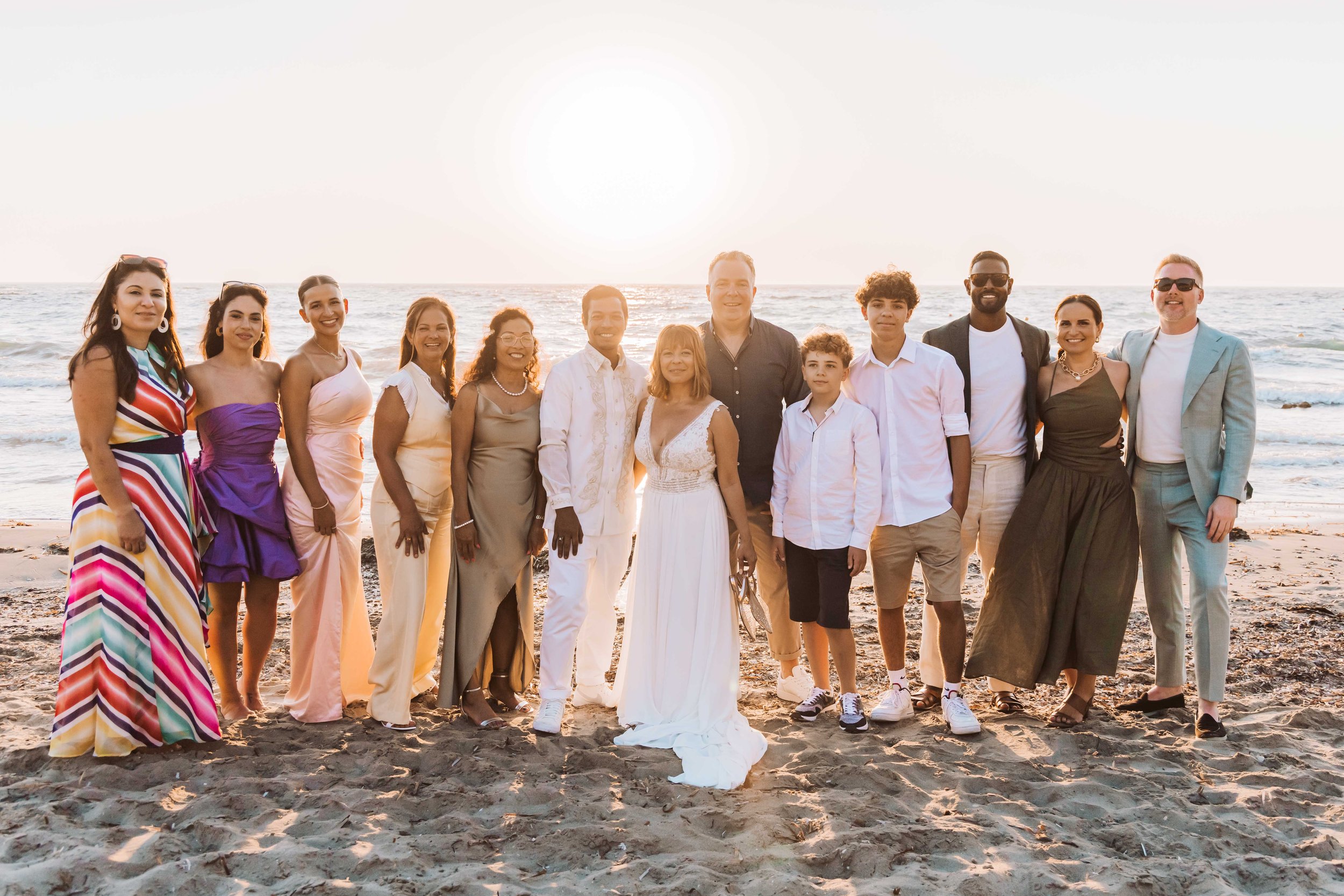 Group of people standing together on a beach during sunset, dressed in semi-formal and casual attire, smiling at the camera.