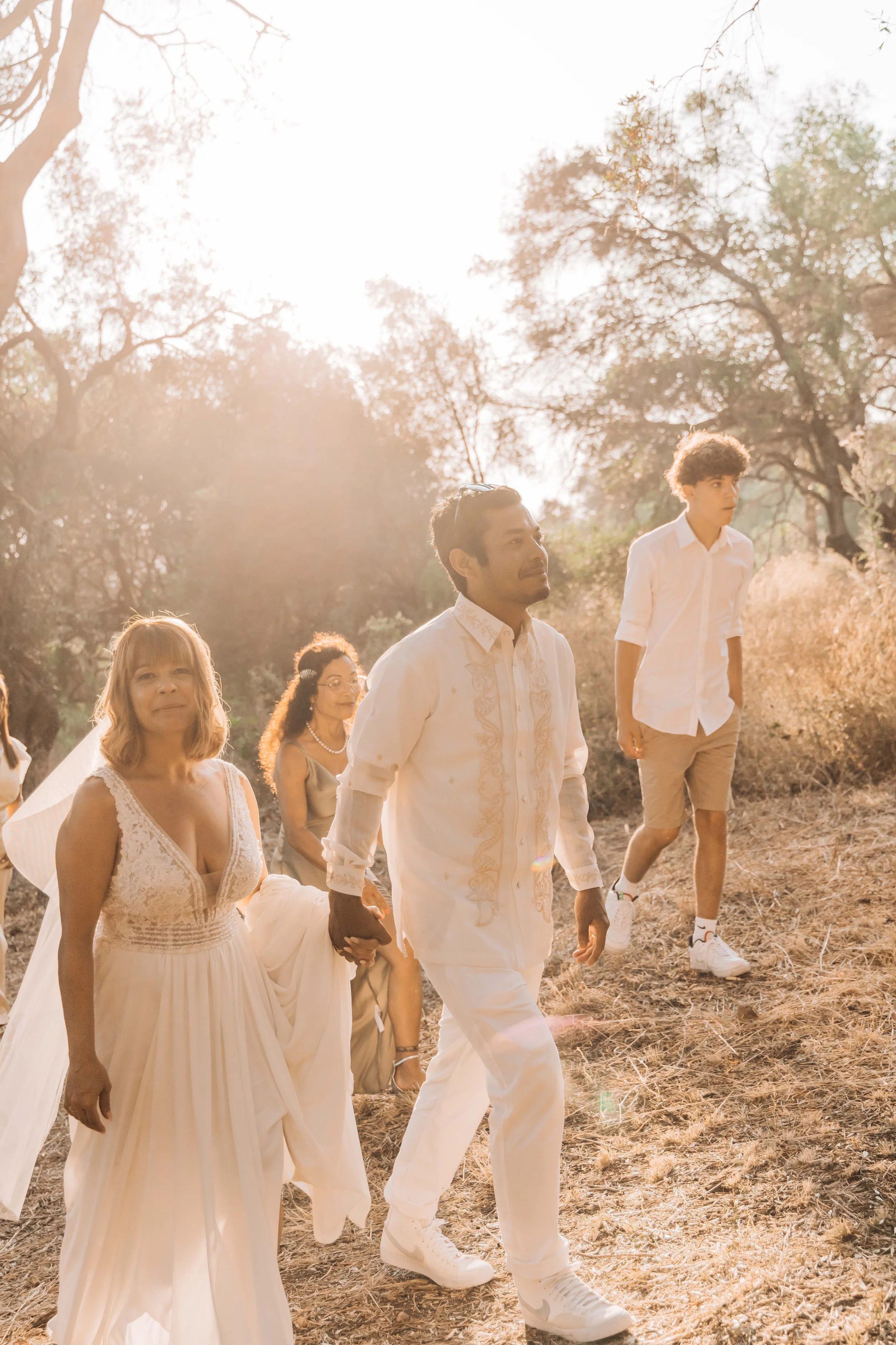 Group of people walking outdoors in a sunny, wooded area, including a woman in a wedding dress, a man in a white embroidered shirt, and a young man in a white shirt and tan shorts.
