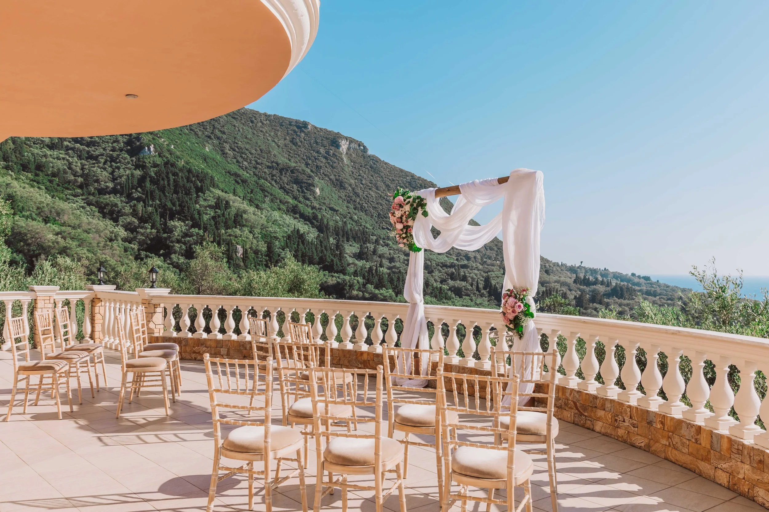 Outdoor wedding ceremony setup on a terrace with a mountain view, featuring a white fabric-draped wedding arch decorated with pink and white flowers, surrounded by white chairs.