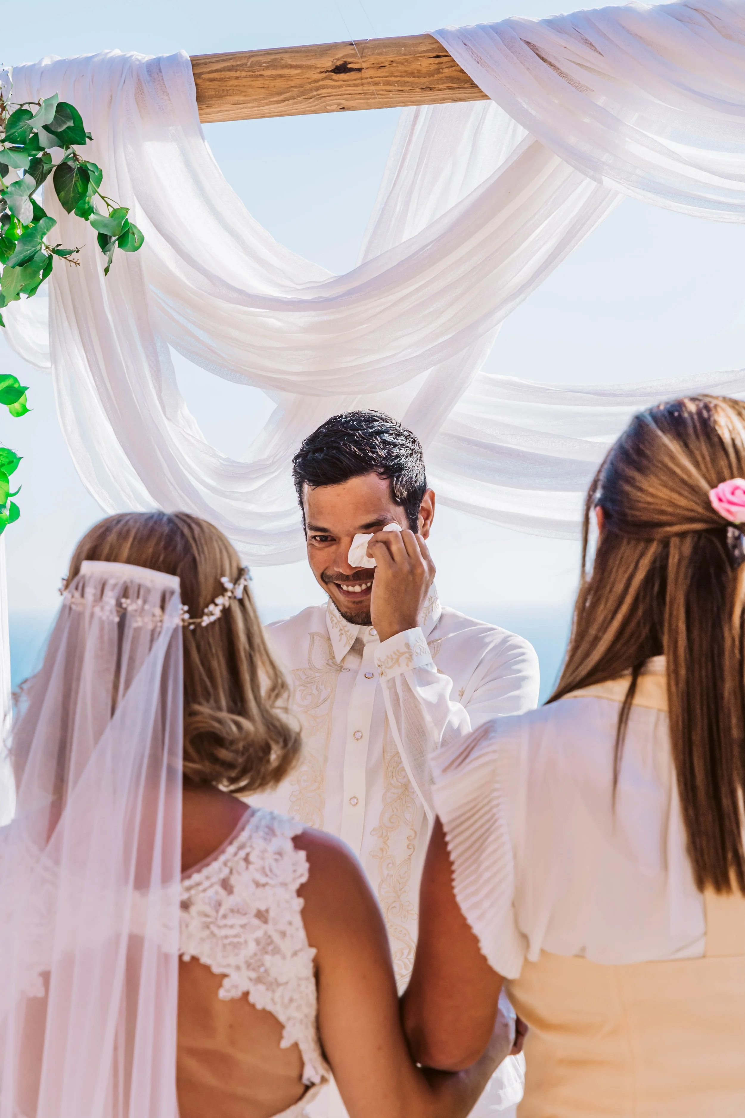 A wedding ceremony by the beach with a groom wiping away a tear, bride in lace dress and veil, and a woman with a flower in her hair, under a white draped canopy.