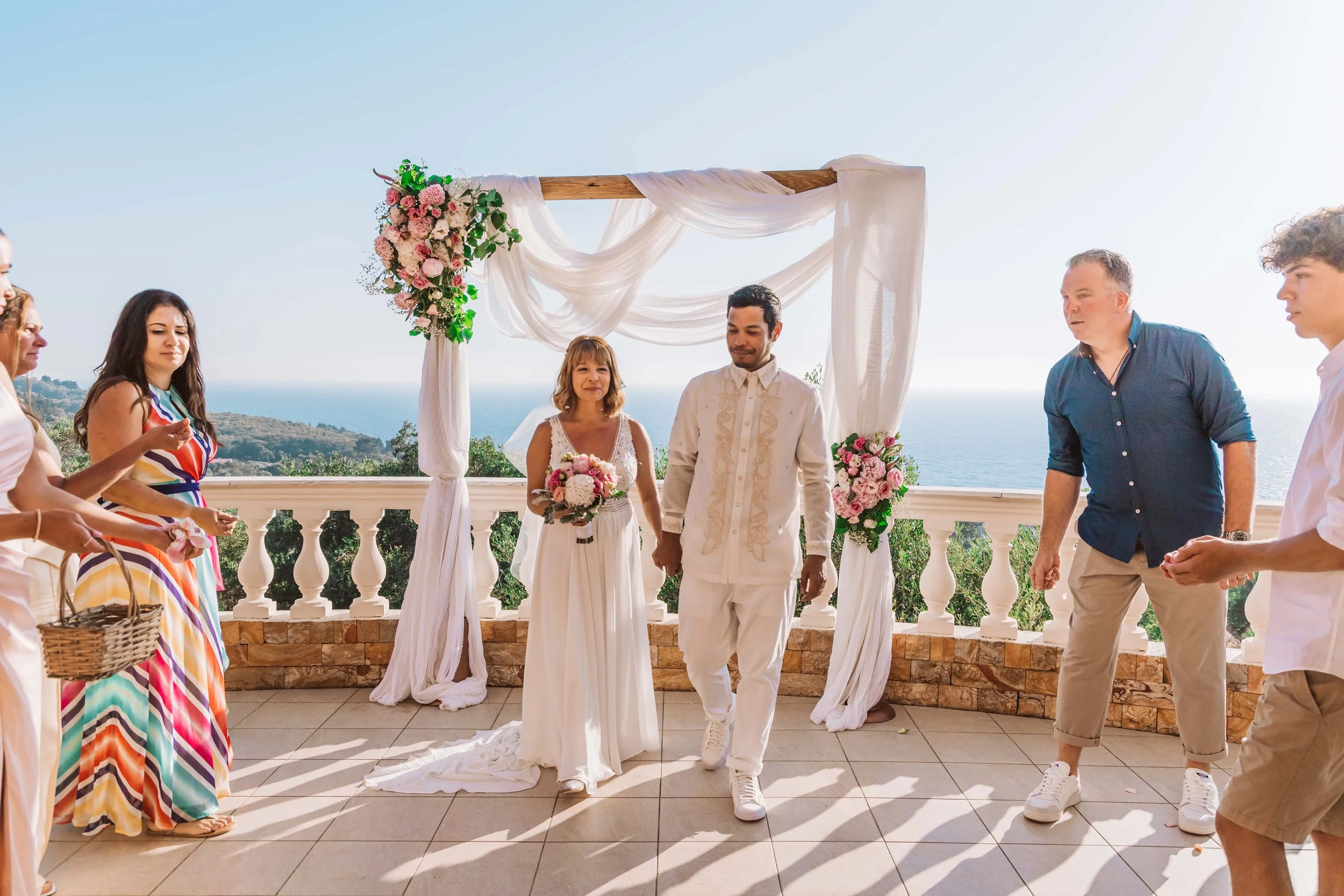 A couple is getting married outdoors under a decorated arch with pink and white flowers, overlooking the ocean, with friends and family around them.