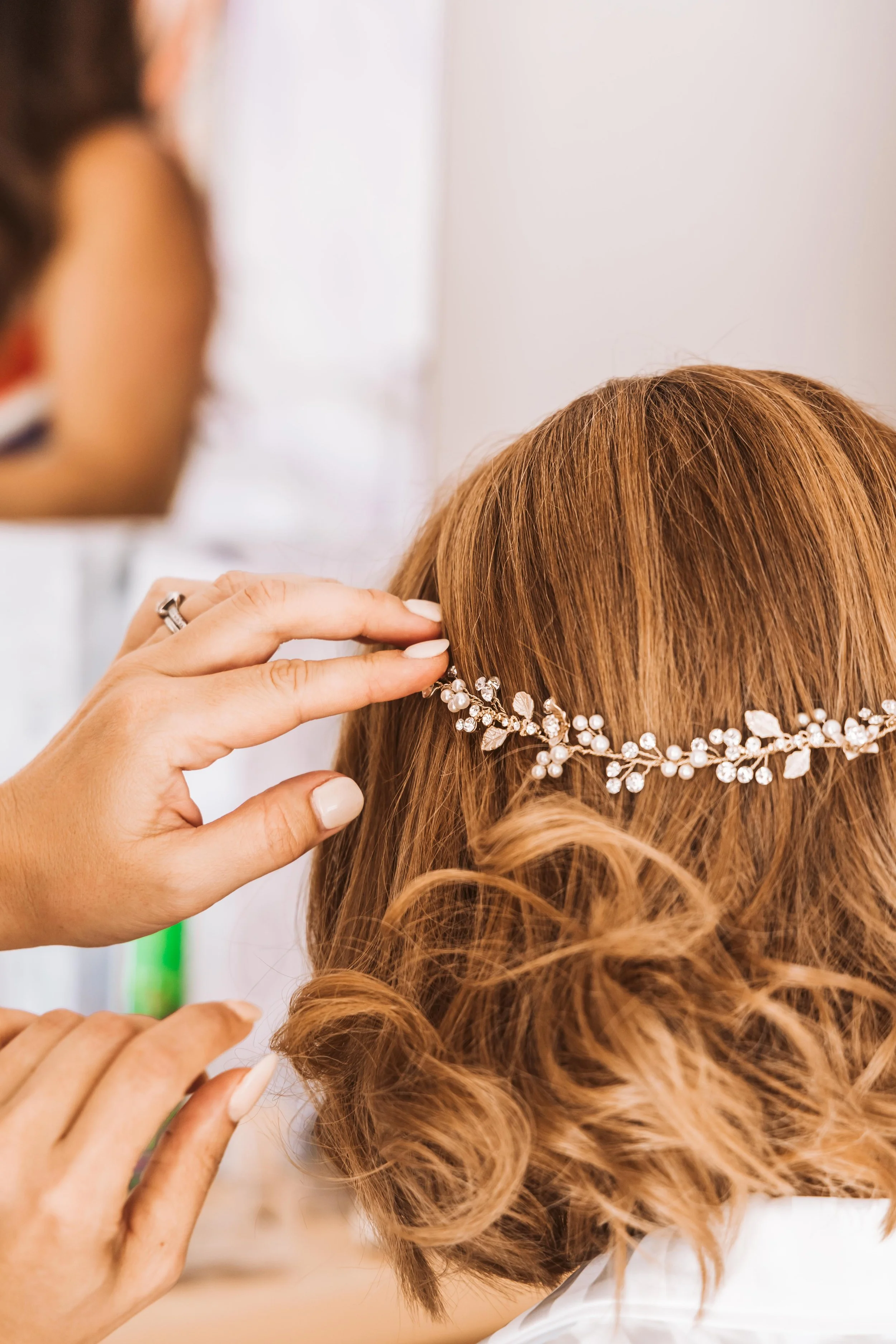 A woman adjusting a decorative pearl and crystal headpiece in her red hair, with another woman in the background.