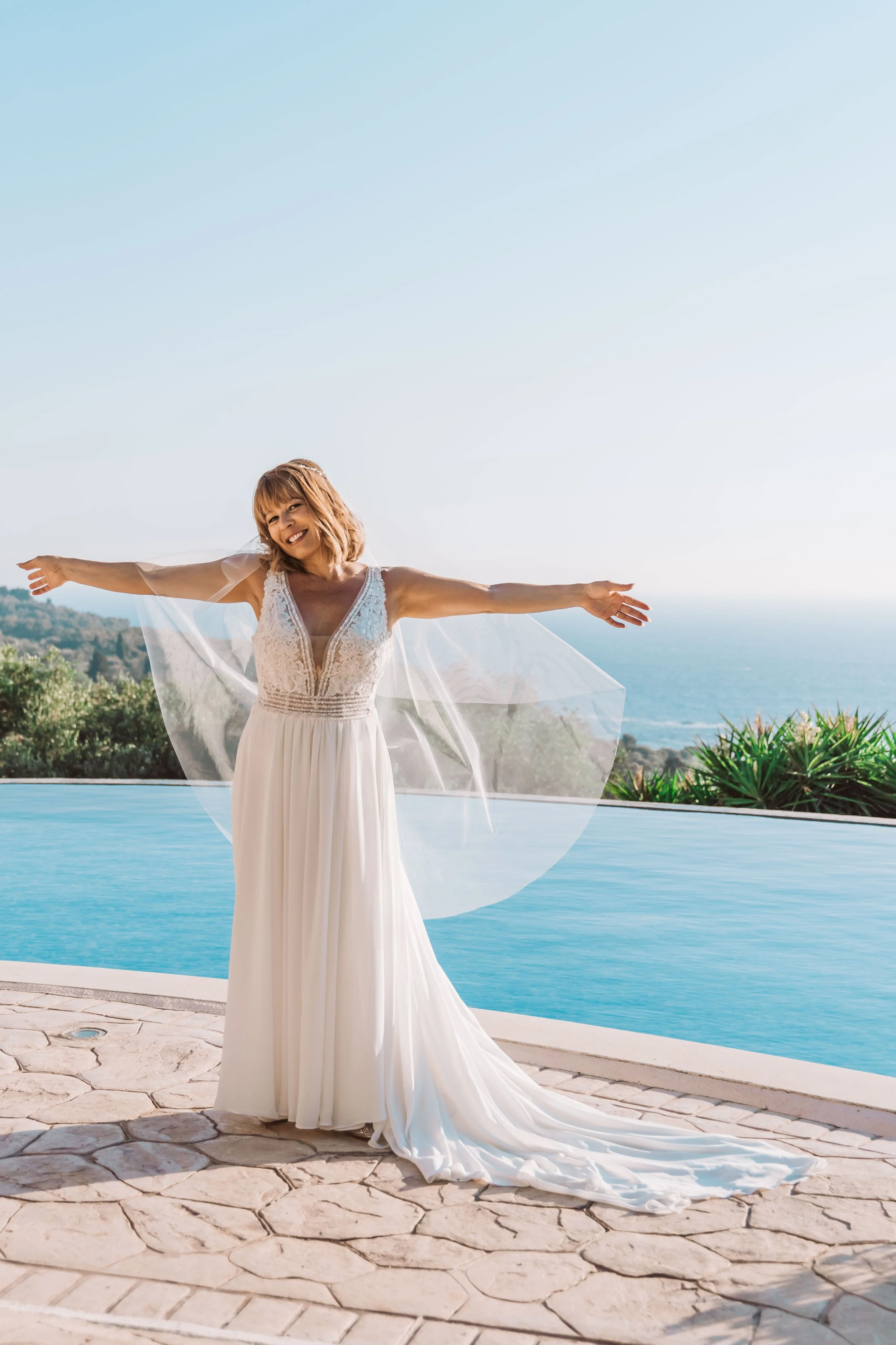 A woman in a white dress standing by a pool with arms outstretched, smiling, with a scenic ocean view in the background.