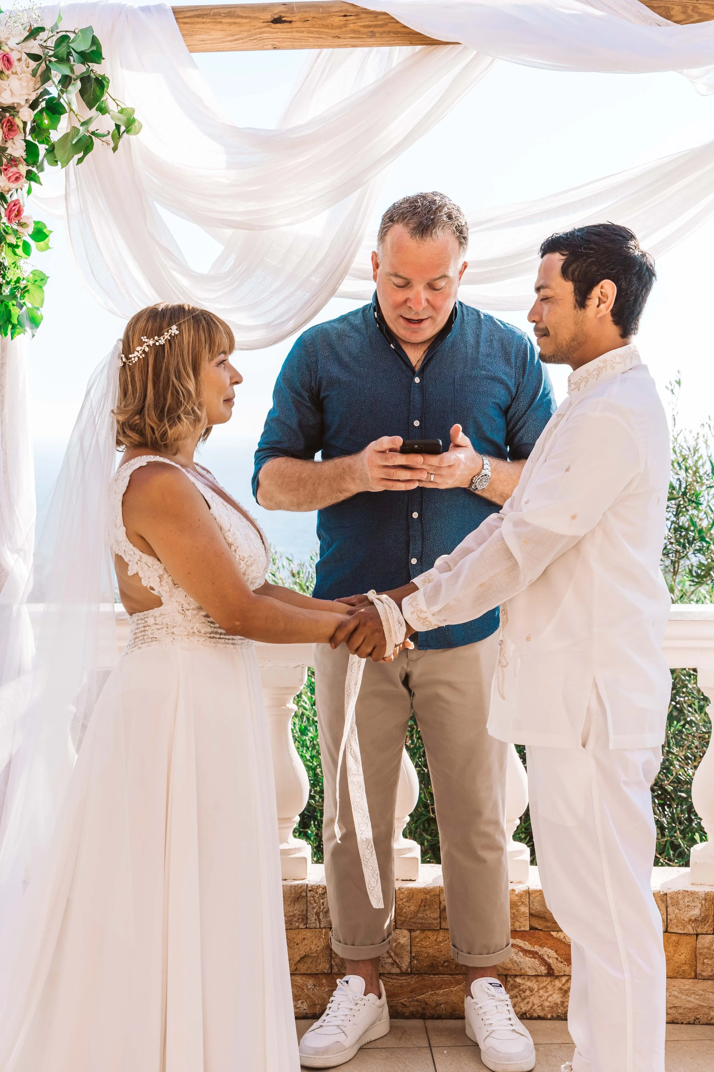 A couple gets married outdoors, holding hands while facing each other and exchanging vows. The officiant stands behind them, looking at a phone. The scene is decorated with flowing white fabric and floral arrangements.