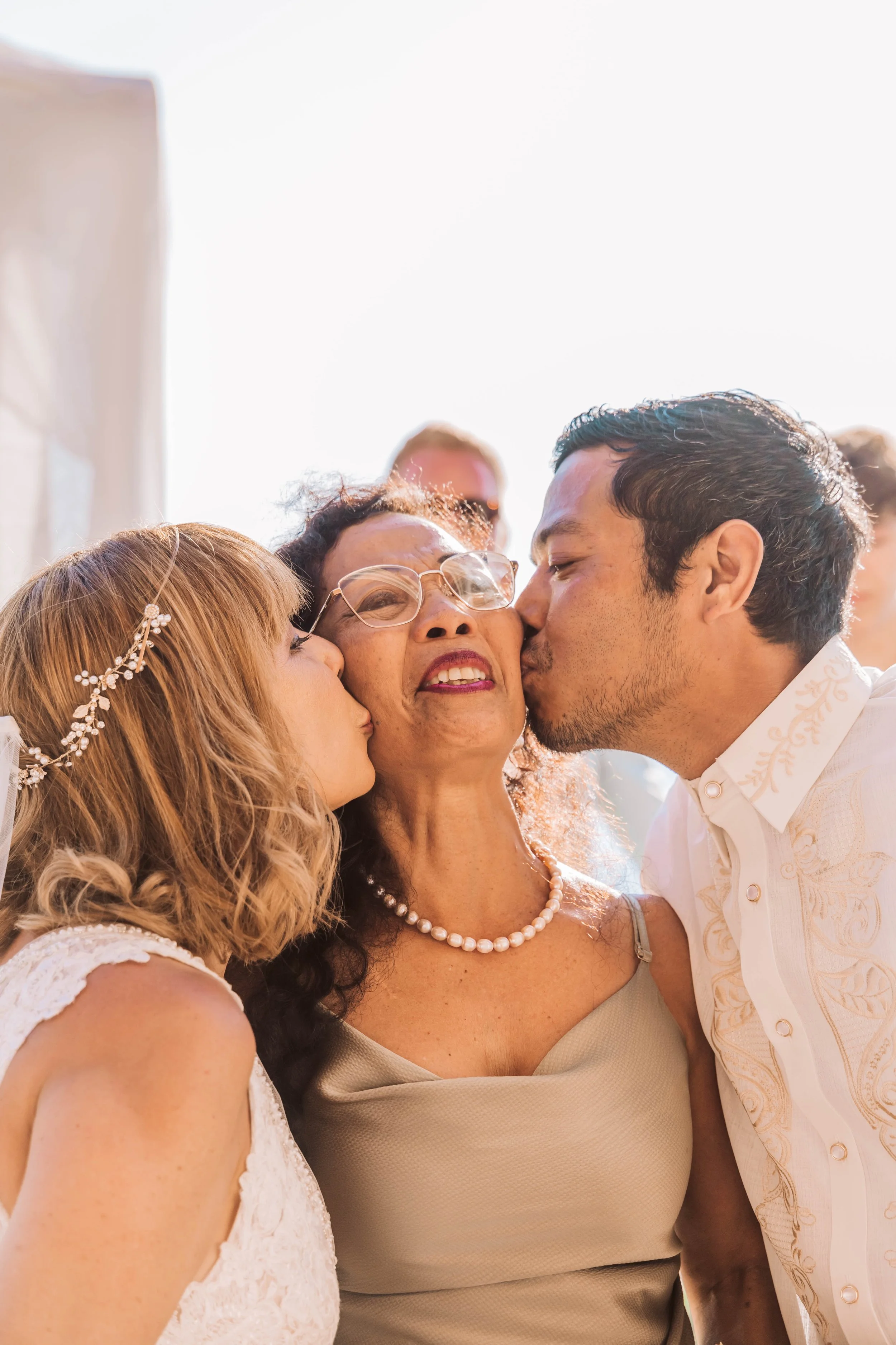 A woman with glasses and a pearl necklace receives kisses on the cheeks from a young woman and a young man at her wedding celebration.