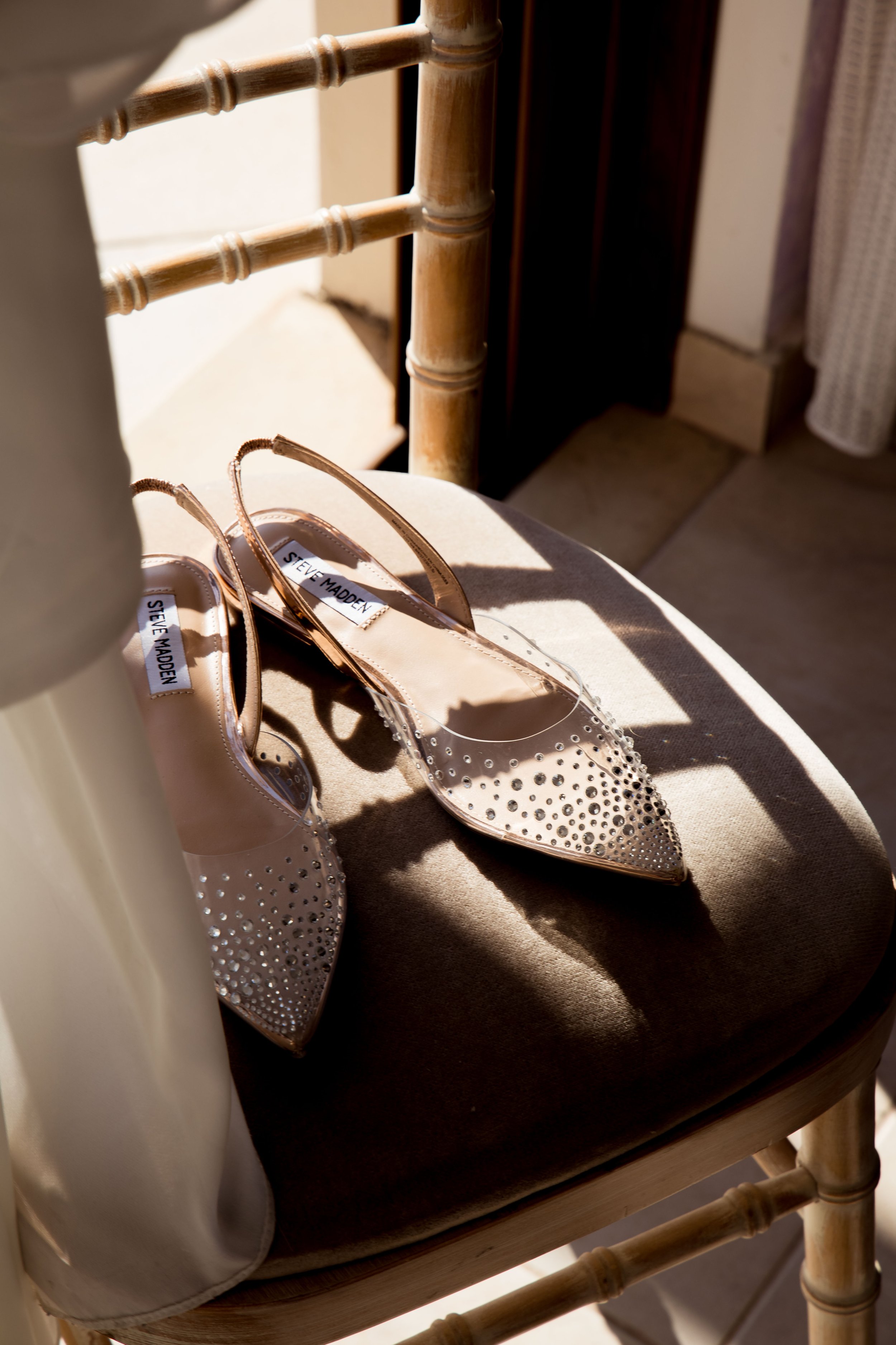 A pair of high-heeled shoes with a transparent body and rhinestone embellishments, placed on a beige cushioned chair near a window with sunlight and shadows.