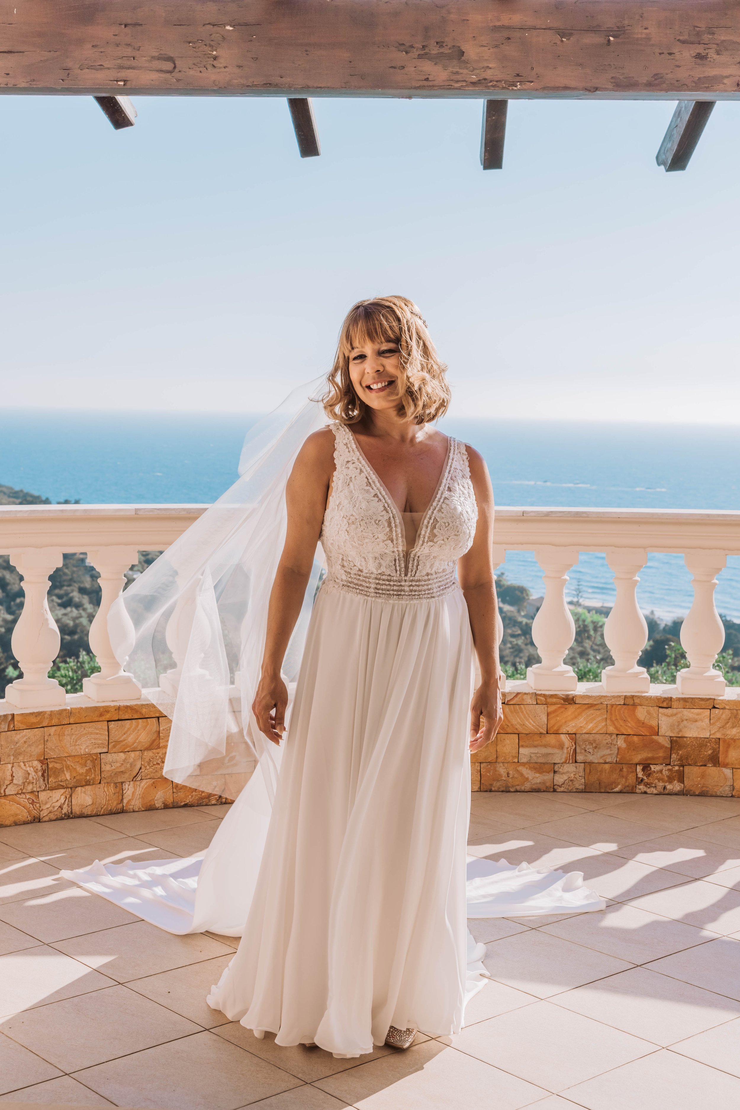 A woman in a white wedding dress and veil stands on a balcony with a view of the ocean and blue sky in the background.