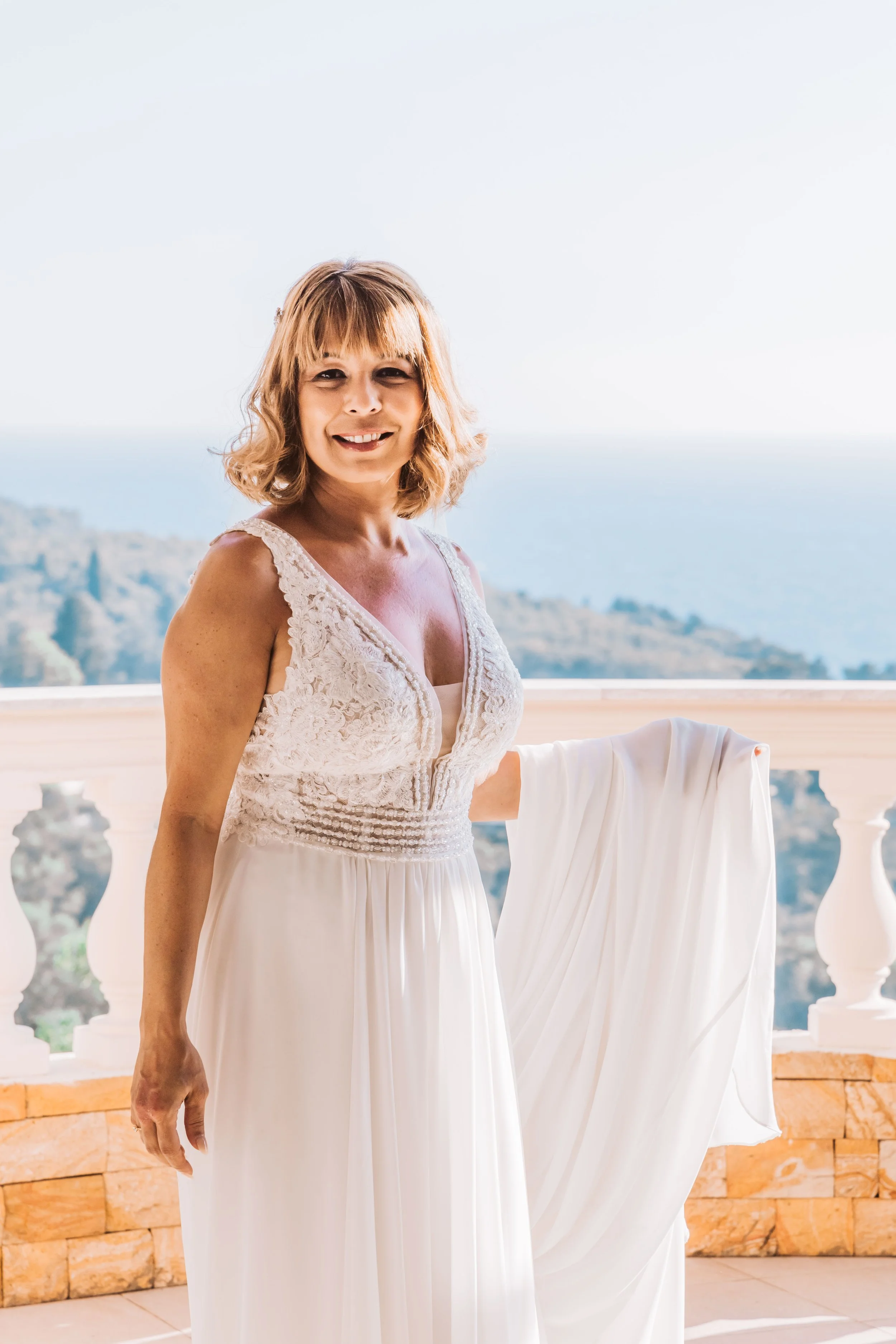 Woman in a white lace dress standing on a balcony with a scenic view of trees, hills, and water in the background, smiling at the camera.