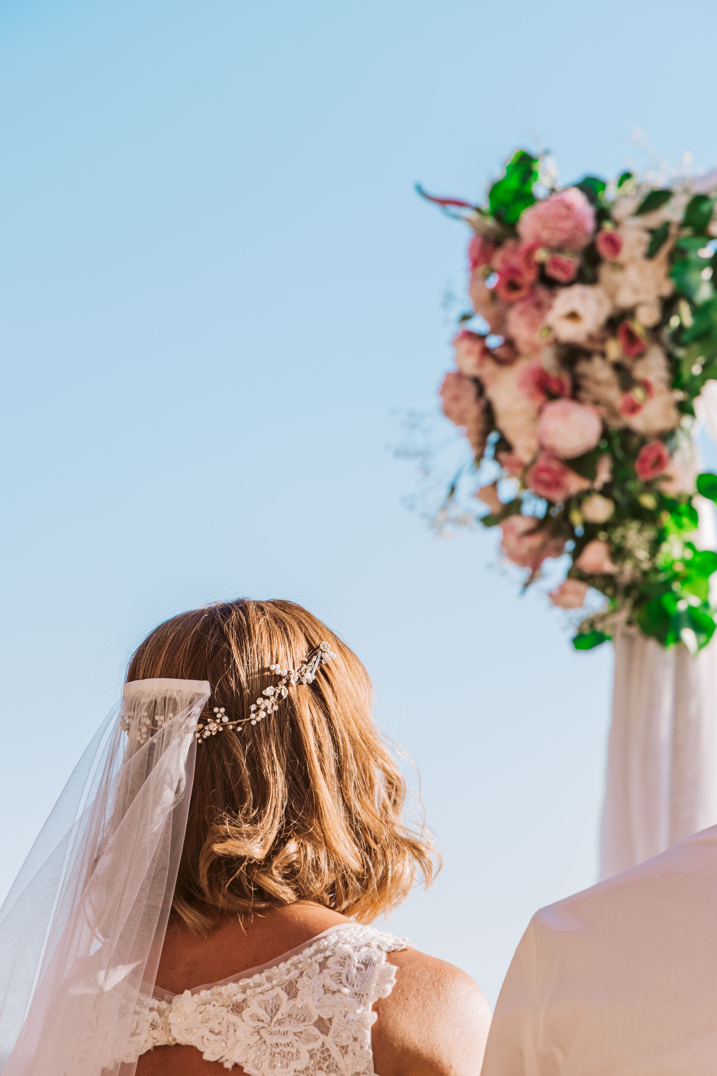 Back view of a bride with wavy blonde hair wearing a lace wedding dress, veil, and decorative headpiece, standing outdoors during a wedding ceremony with a large floral arrangement in the background against a clear blue sky.