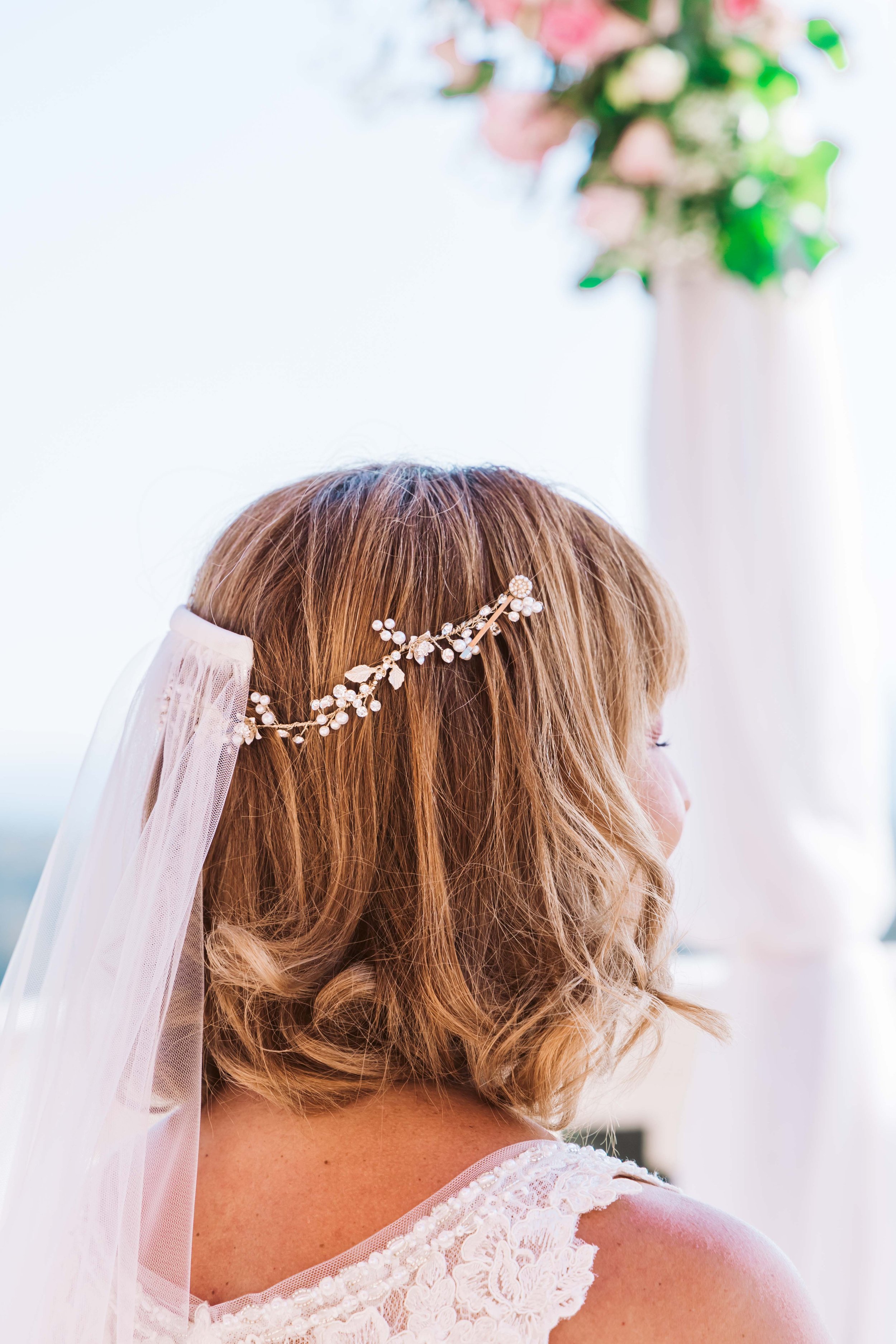 Close-up of a bride with medium-length, wavy, light brown hair, wearing a floral hair accessory and veil, against a bright background with a blurred floral arrangement.