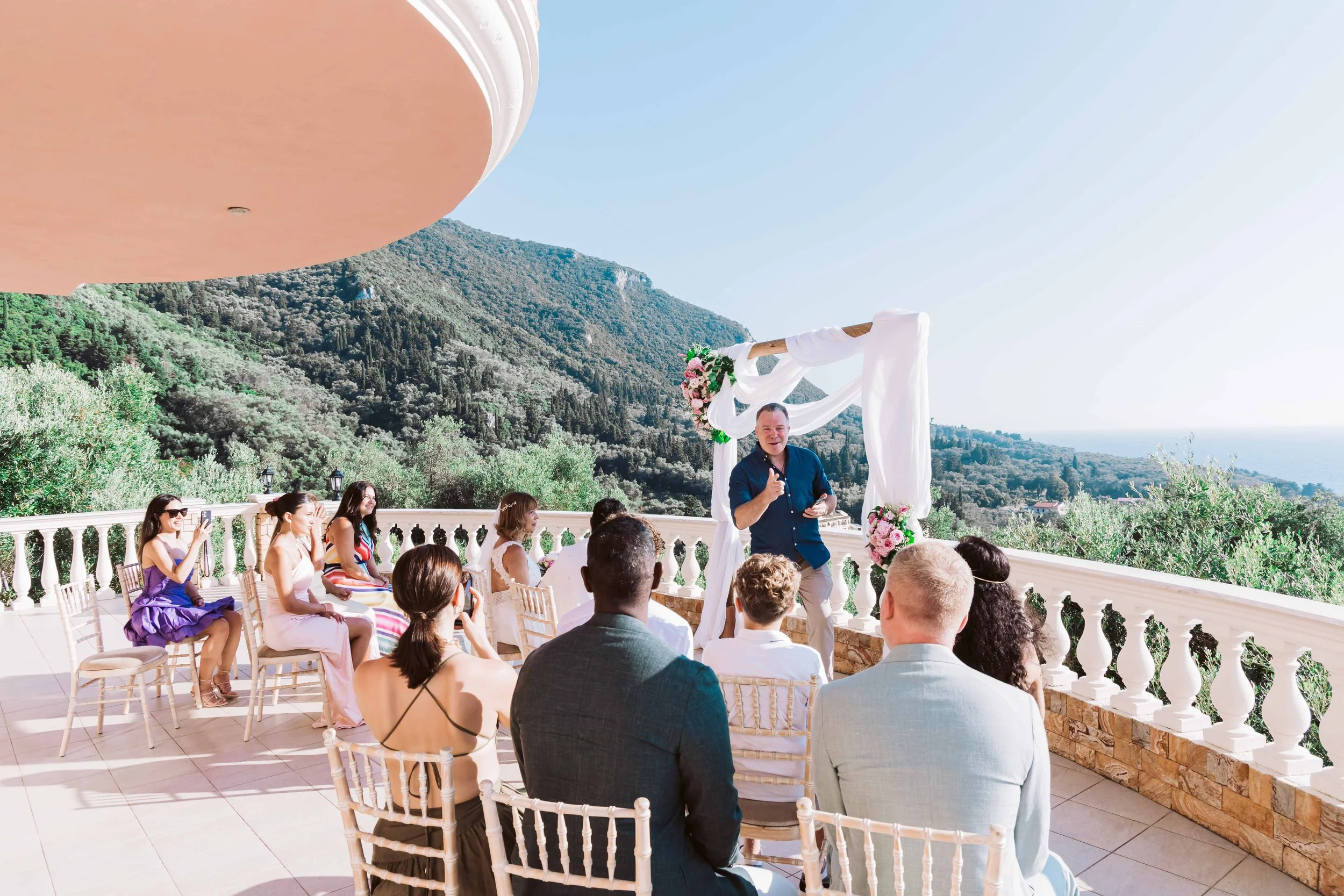 A wedding ceremony taking place on a balcony with a view of lush green mountains and the ocean. The officiant is speaking, and guests are seated, some taking photos, while the decorated arch with pink flowers stands prominently.