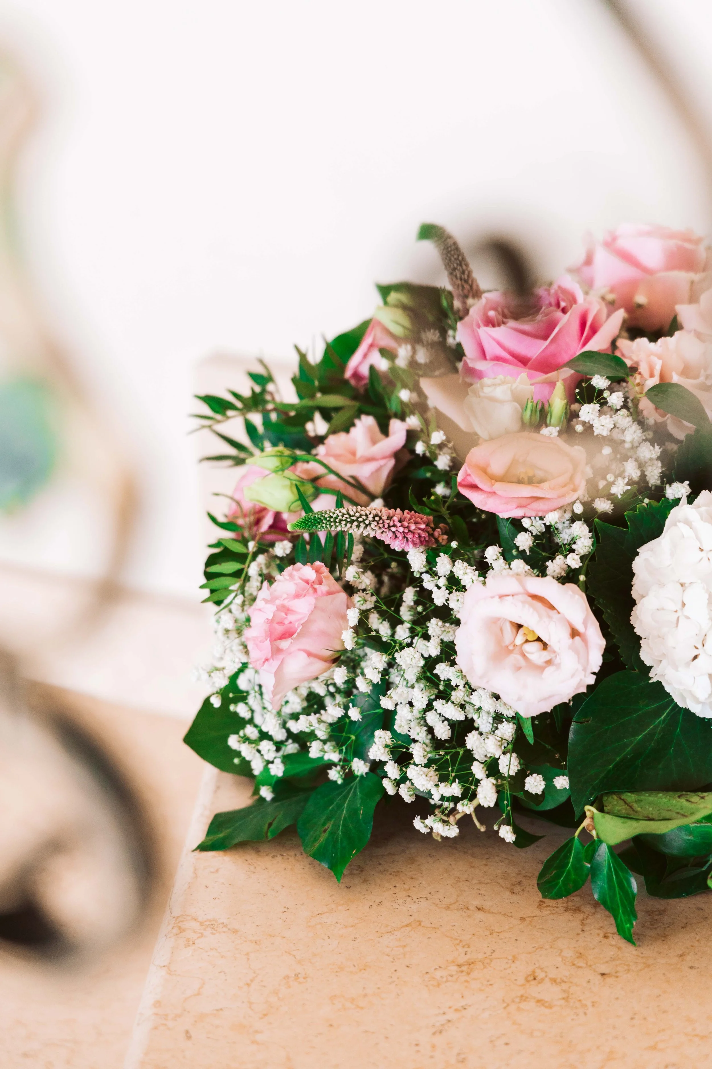A bouquet of pink and white flowers, including roses, baby's breath, and greenery, resting on a beige marble surface.