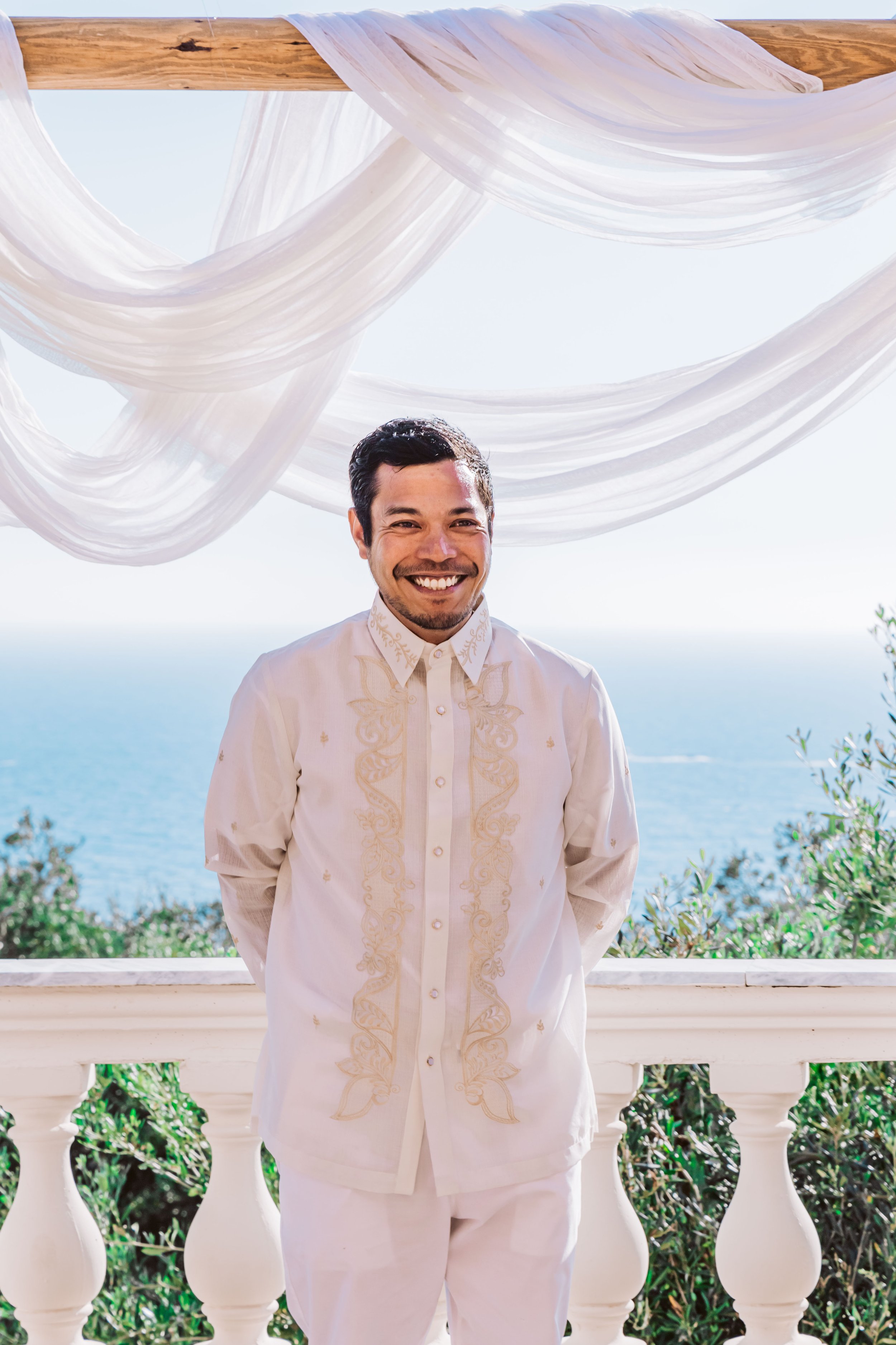 A man smiling in traditional Filipino attire standing on a balcony with ocean view, decorated with white drapes.