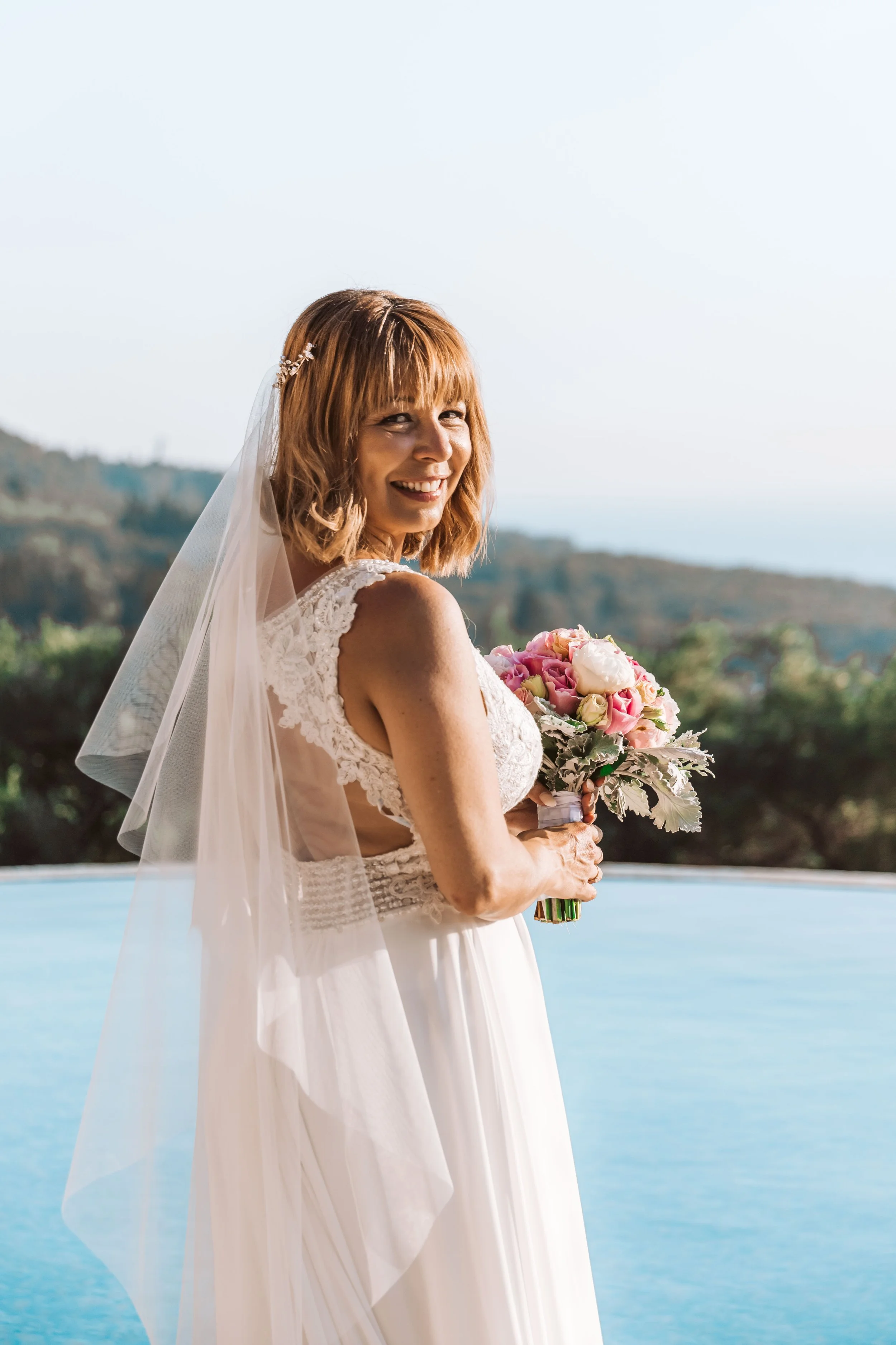 Smiling woman in wedding dress holding a bouquet of pink and white flowers outdoors with a scenic landscape in the background.