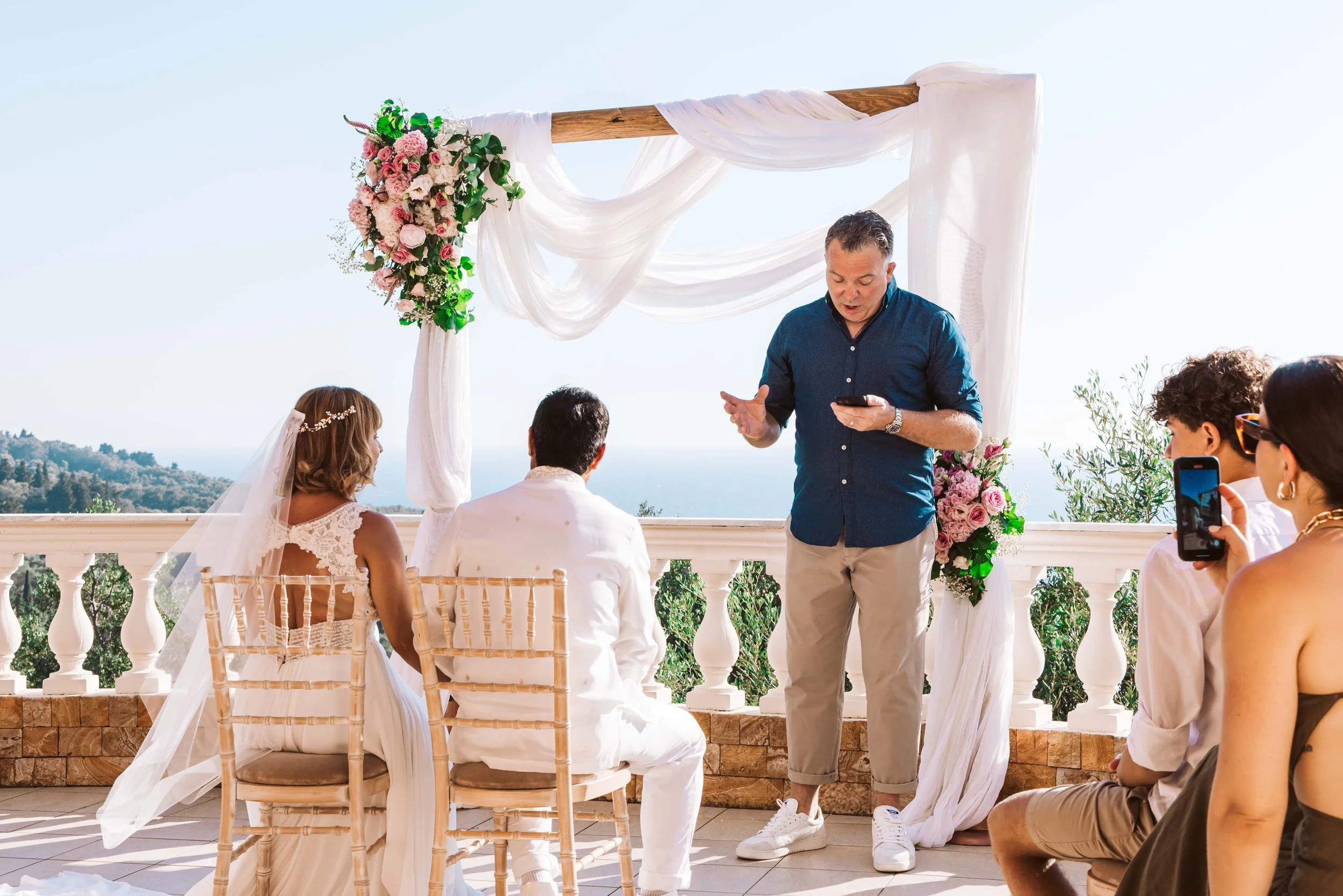 Man officiating a wedding ceremony outdoors on a scenic balcony with an ocean view, placing a ring on a woman's finger, while guests watch and take photos.