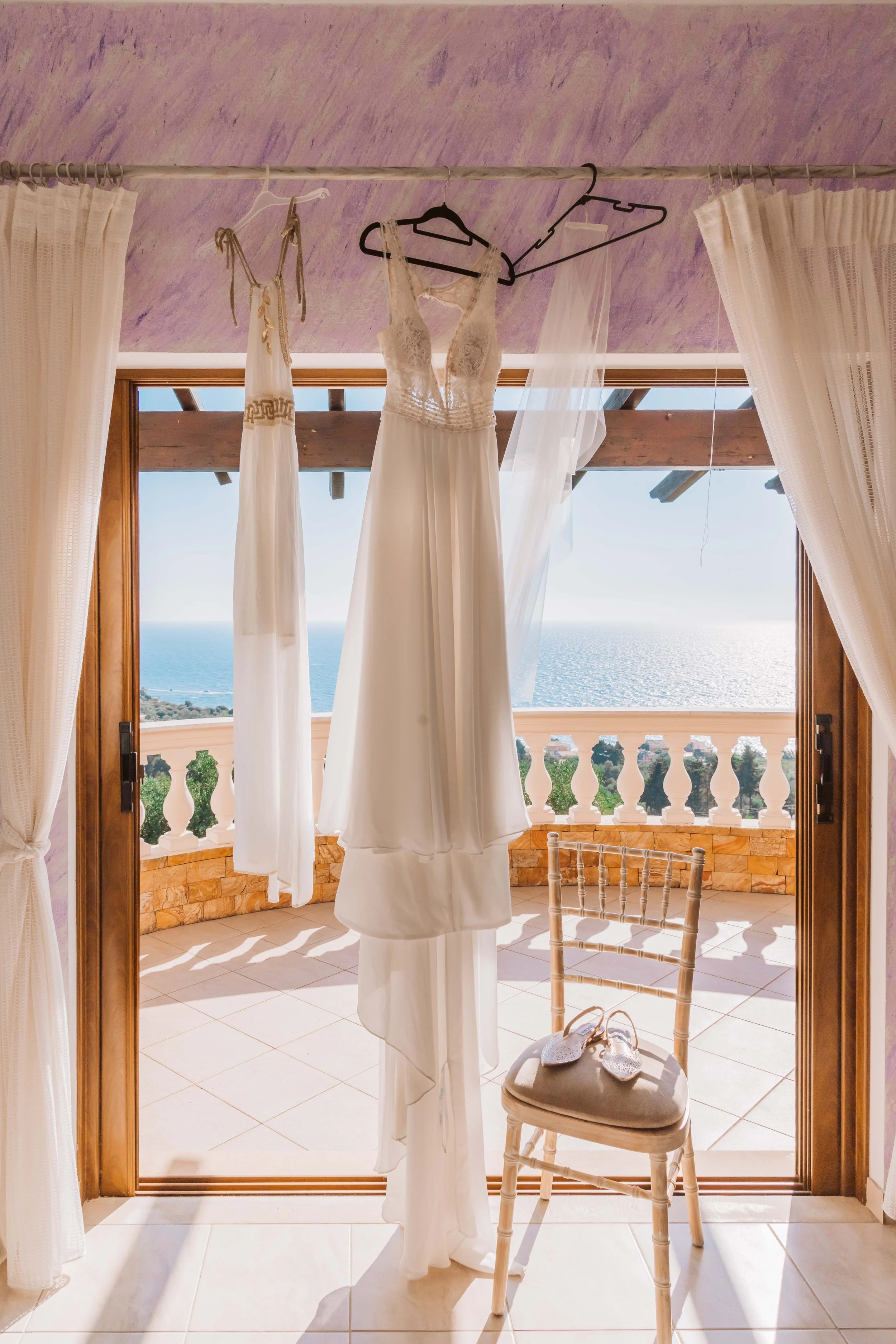 Wedding dresses hanging on hangers in front of a large window with a sea view, a wooden balcony, and a chair with shoes on it.