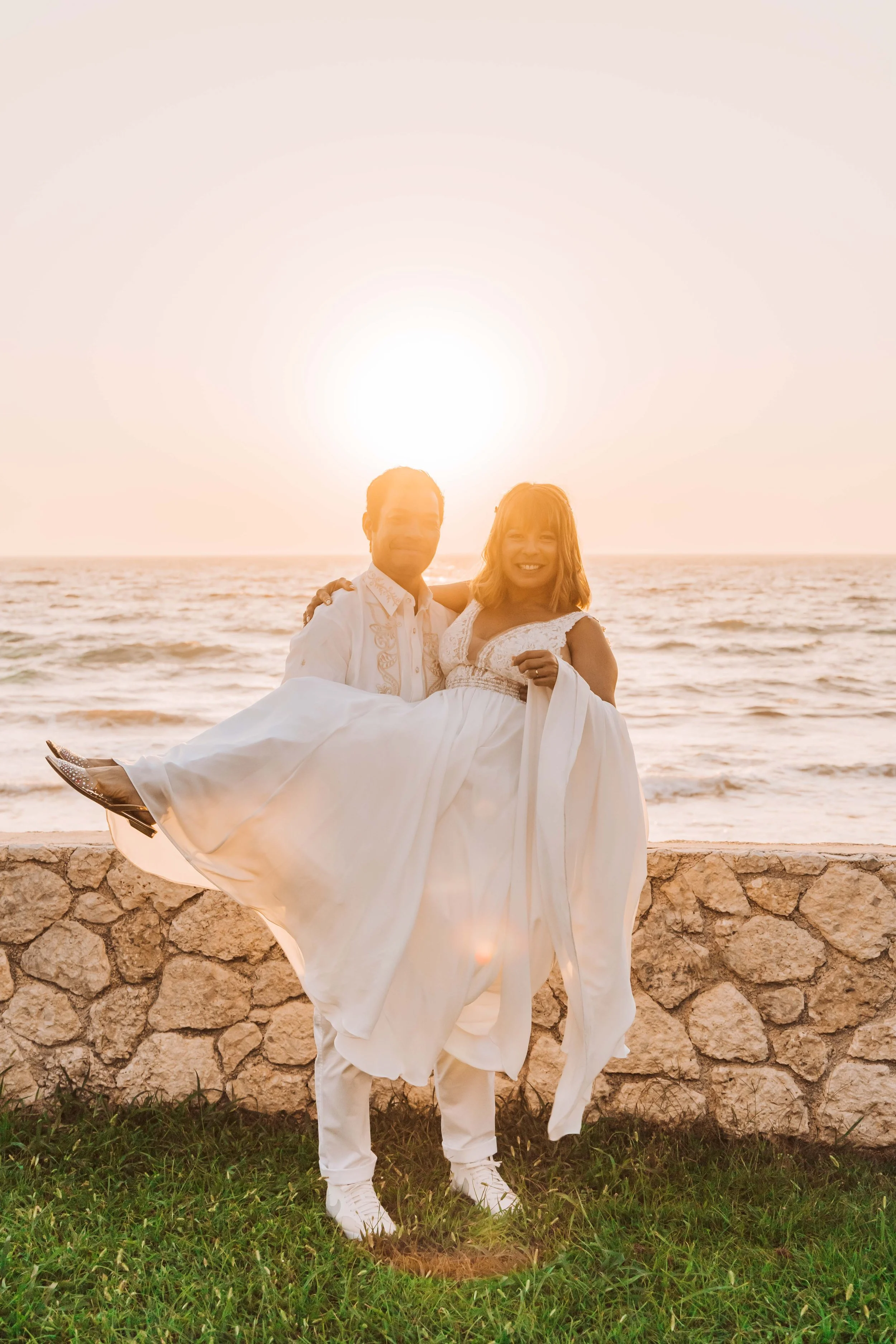 A couple in white wedding attire at the beach during sunset, with the man lifting the woman in his arms.