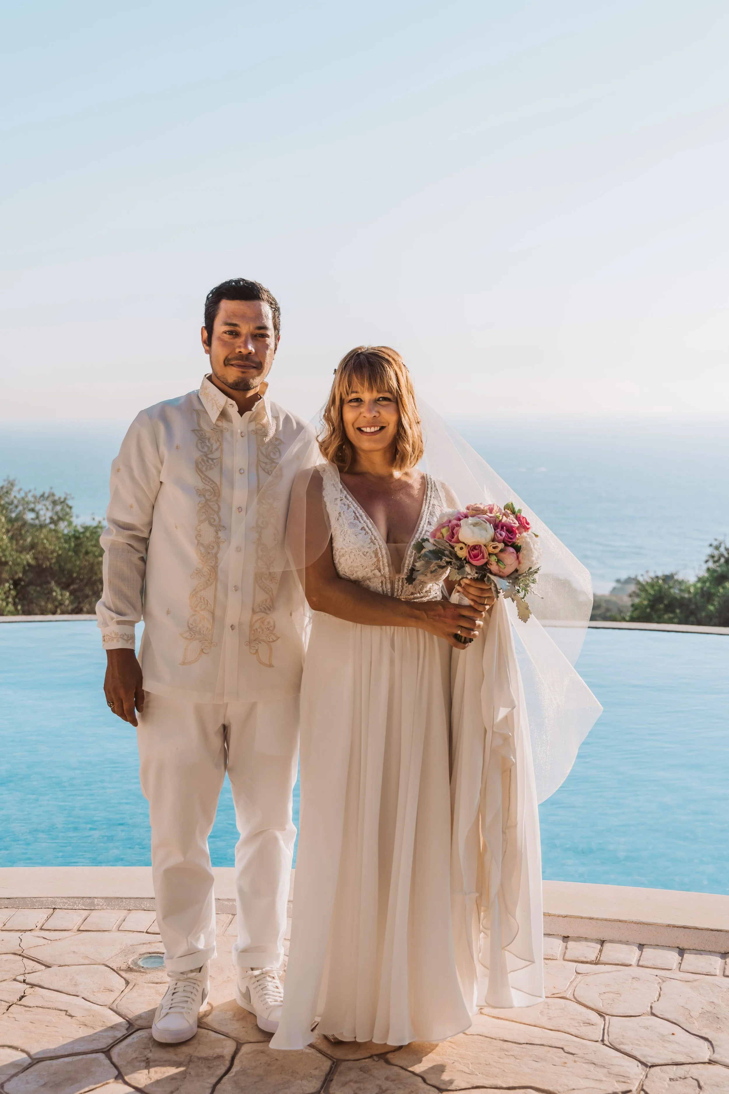 A couple in wedding attire standing outdoors by a pool with the ocean in the background, holding a bouquet of pink, white, and purple flowers, smiling at the camera.