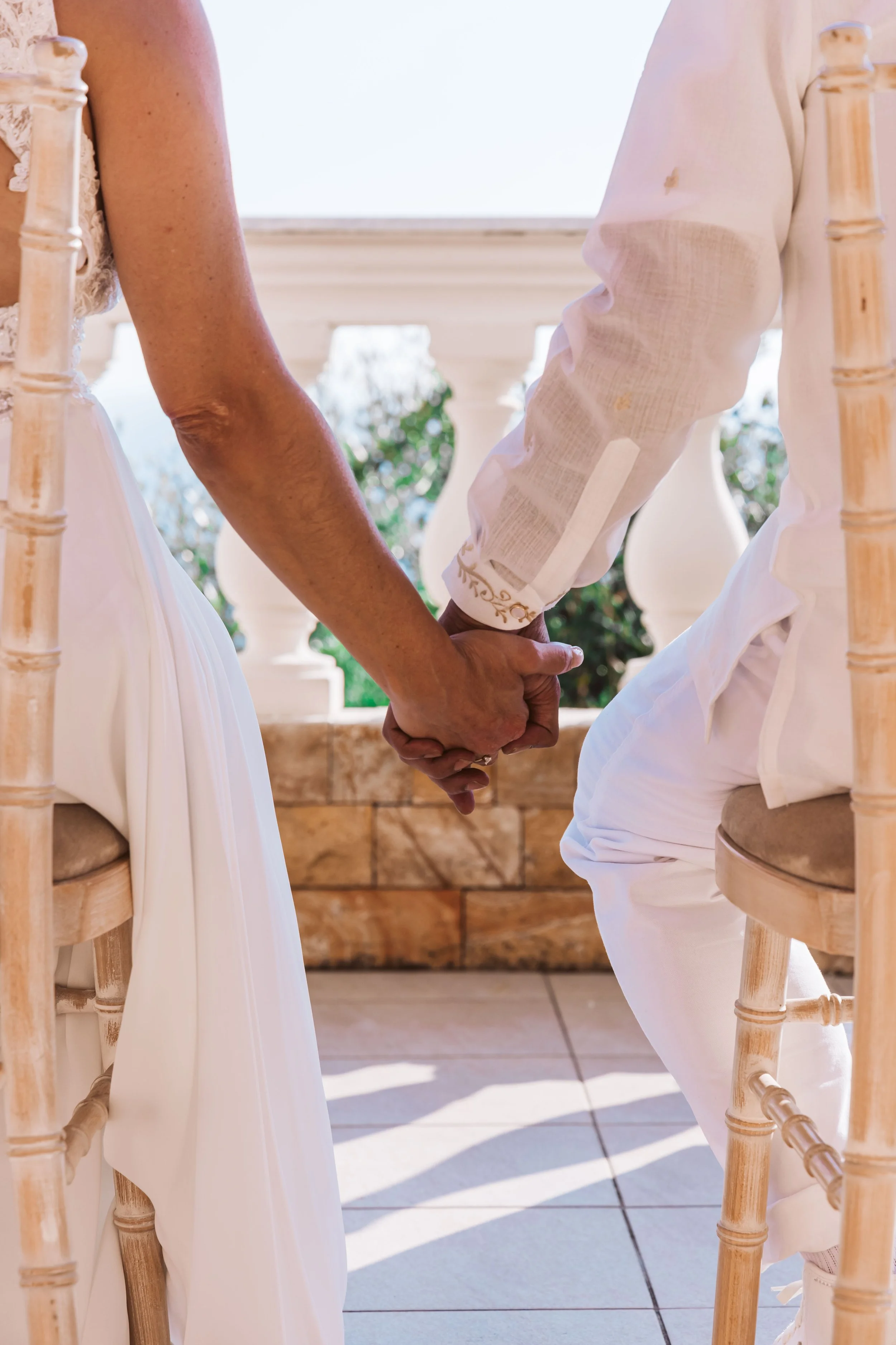 A couple holding hands during a wedding ceremony, sitting on chairs outdoors with sunlight and greenery in the background.