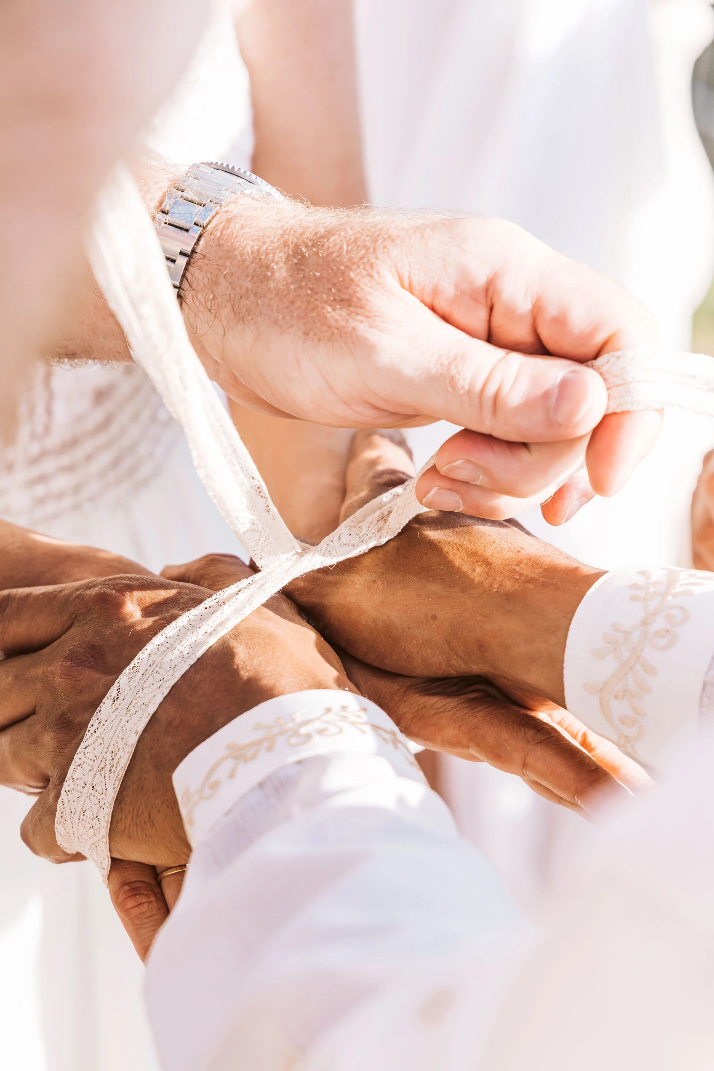 Two people hold hands, one with dark skin and one with light skin, tying white lace ribbons together.