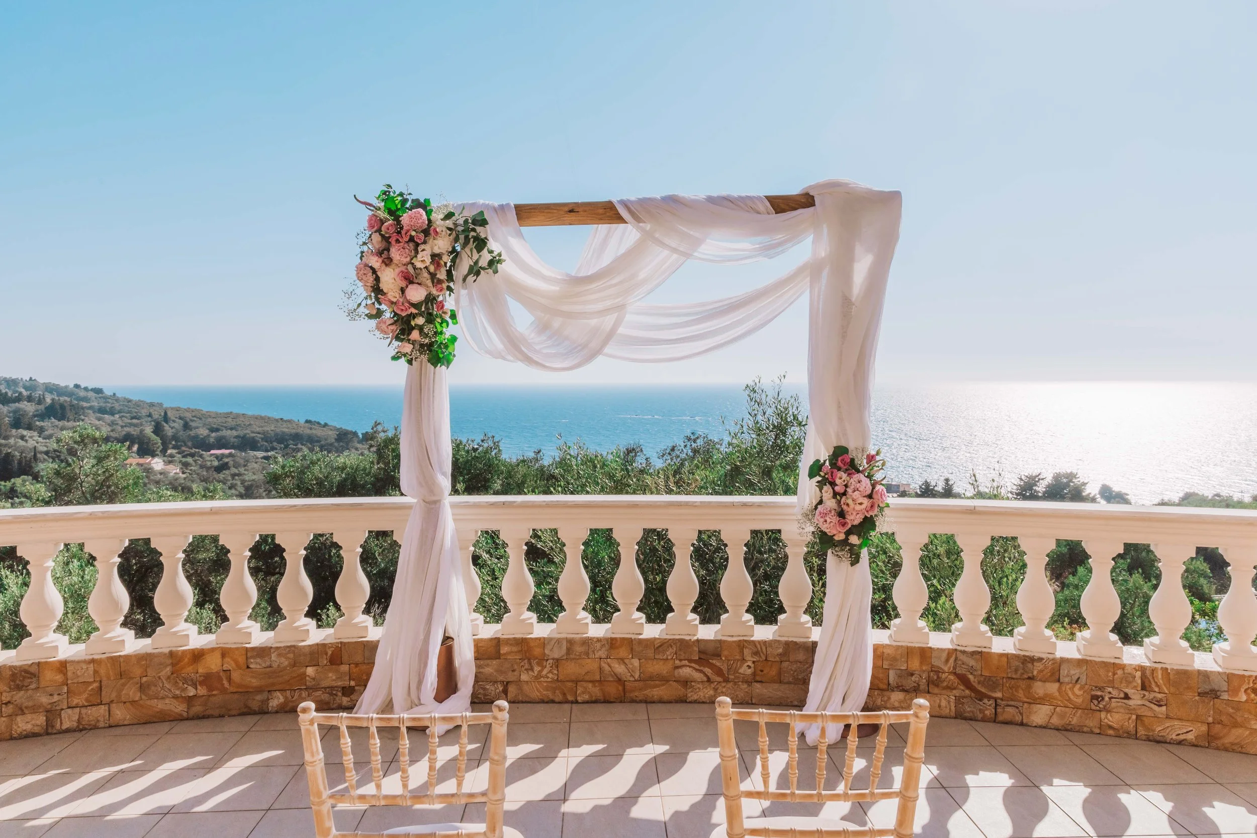 Wedding arch decorated with pink and white flowers, draped with white fabric, overlooking the ocean from a balcony with white balustrades.