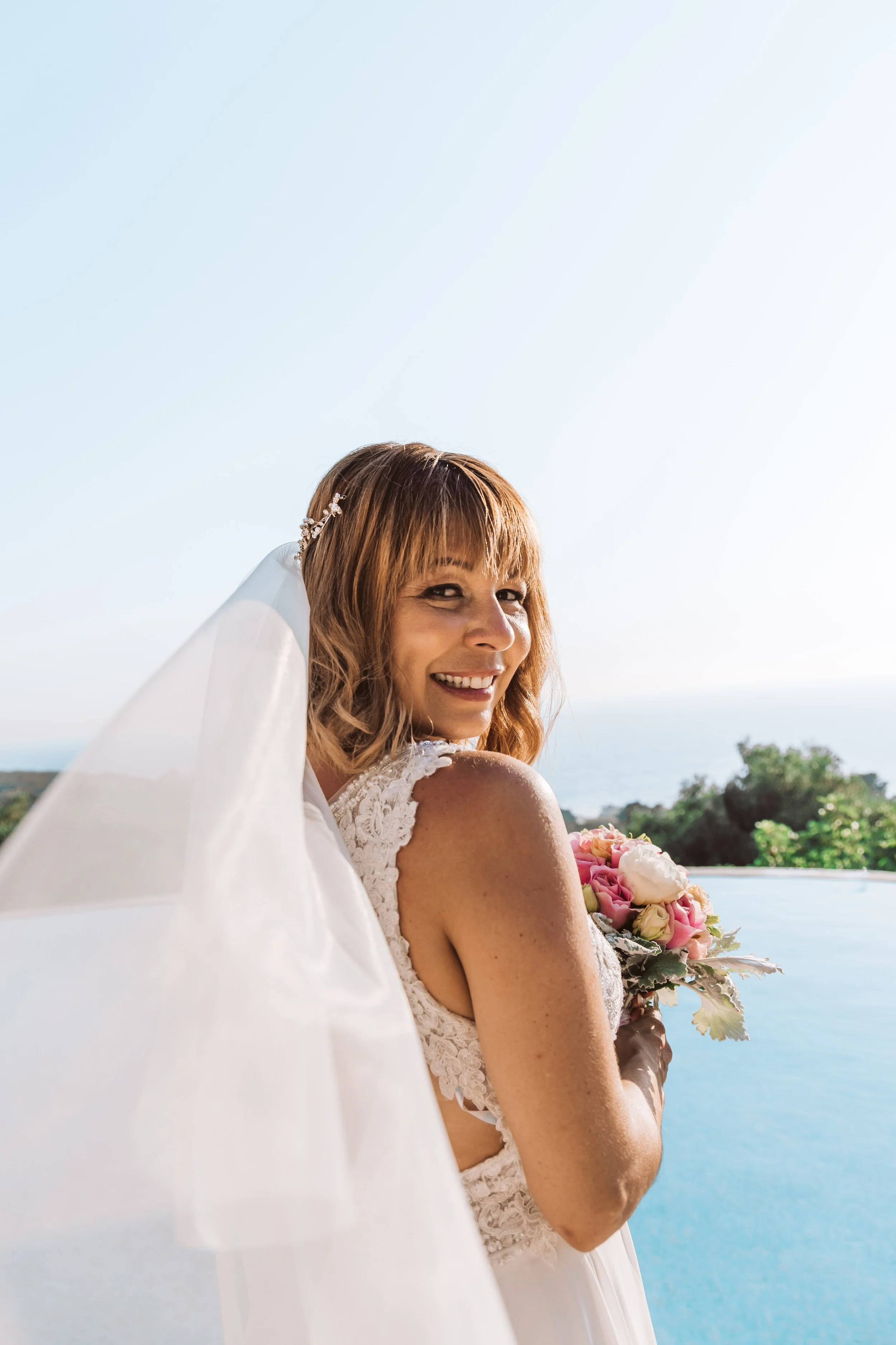 Smiling bride in a white lace wedding dress holding a bouquet of pink and white roses, standing outdoors with a landscape and water in the background.