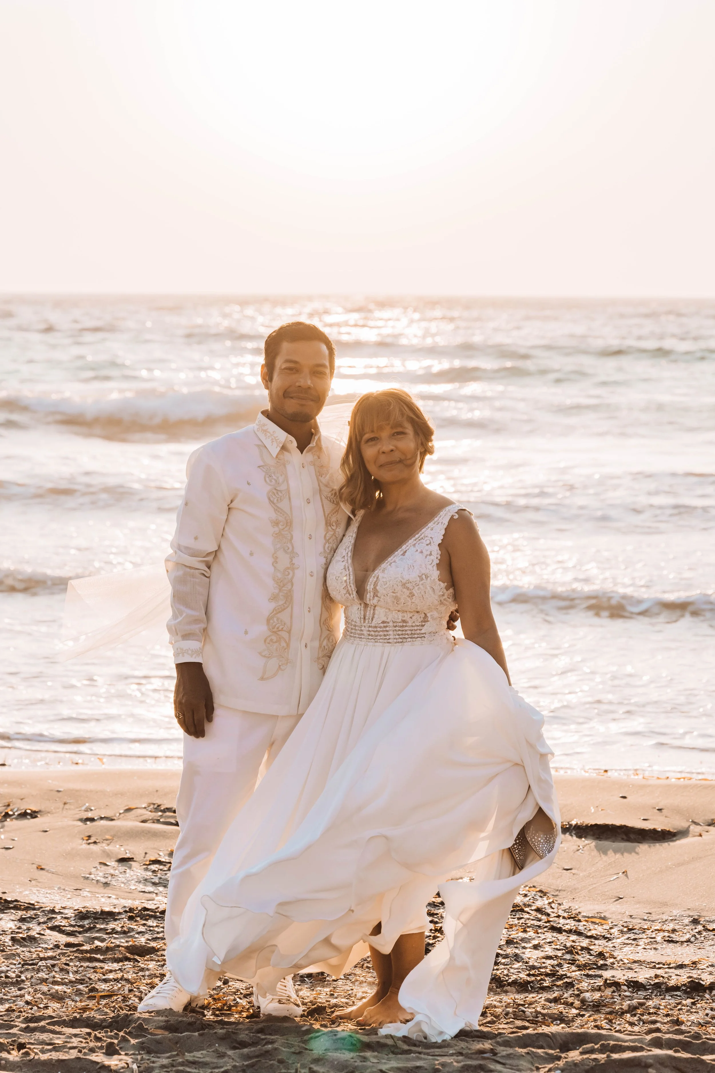 A newlywed couple dressed in white wedding attire standing on the beach at sunset, with the ocean waves in the background.