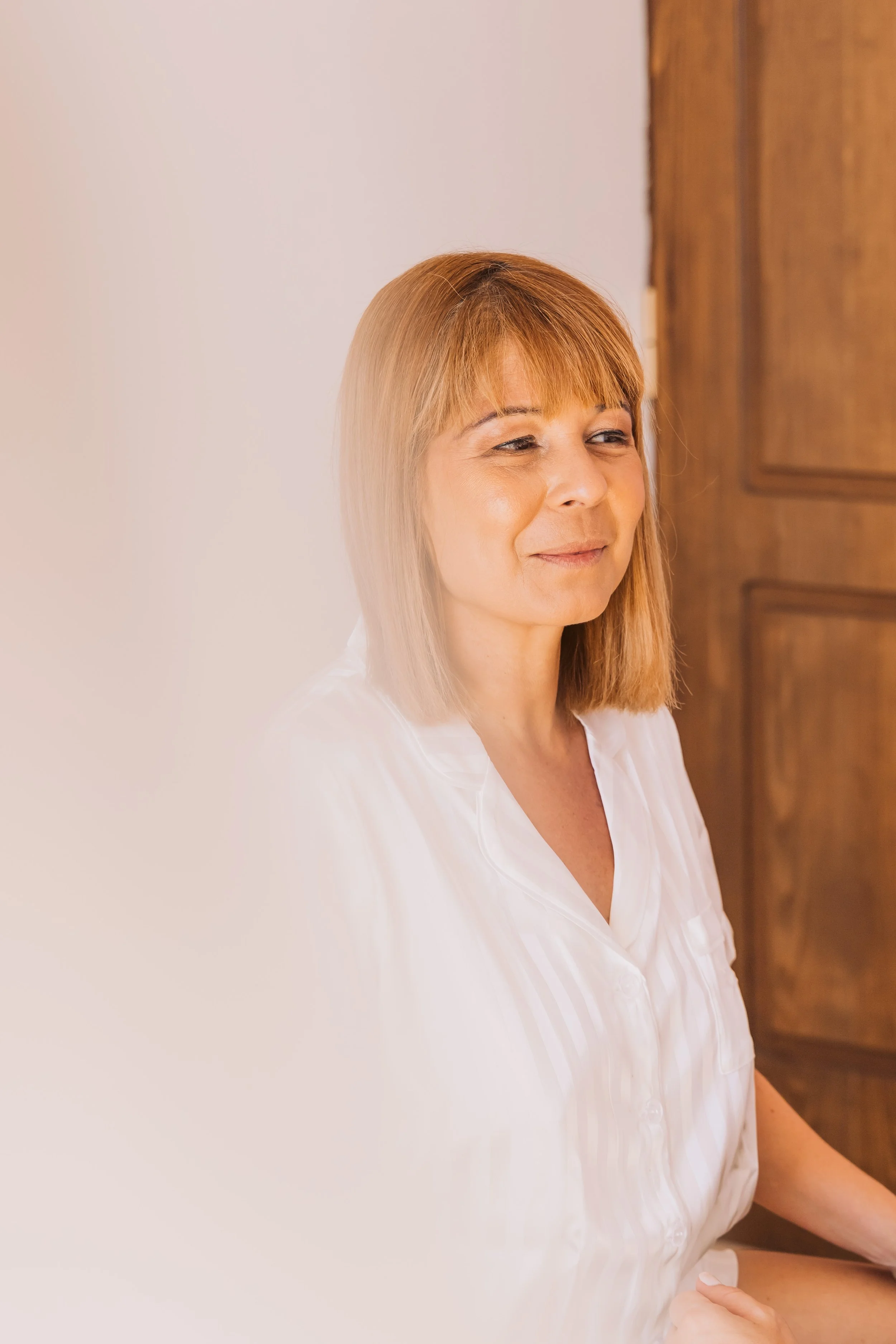 A woman with shoulder-length red hair, wearing a white shirt, standing indoors next to a wooden door, looking slightly to the side with a gentle smile.