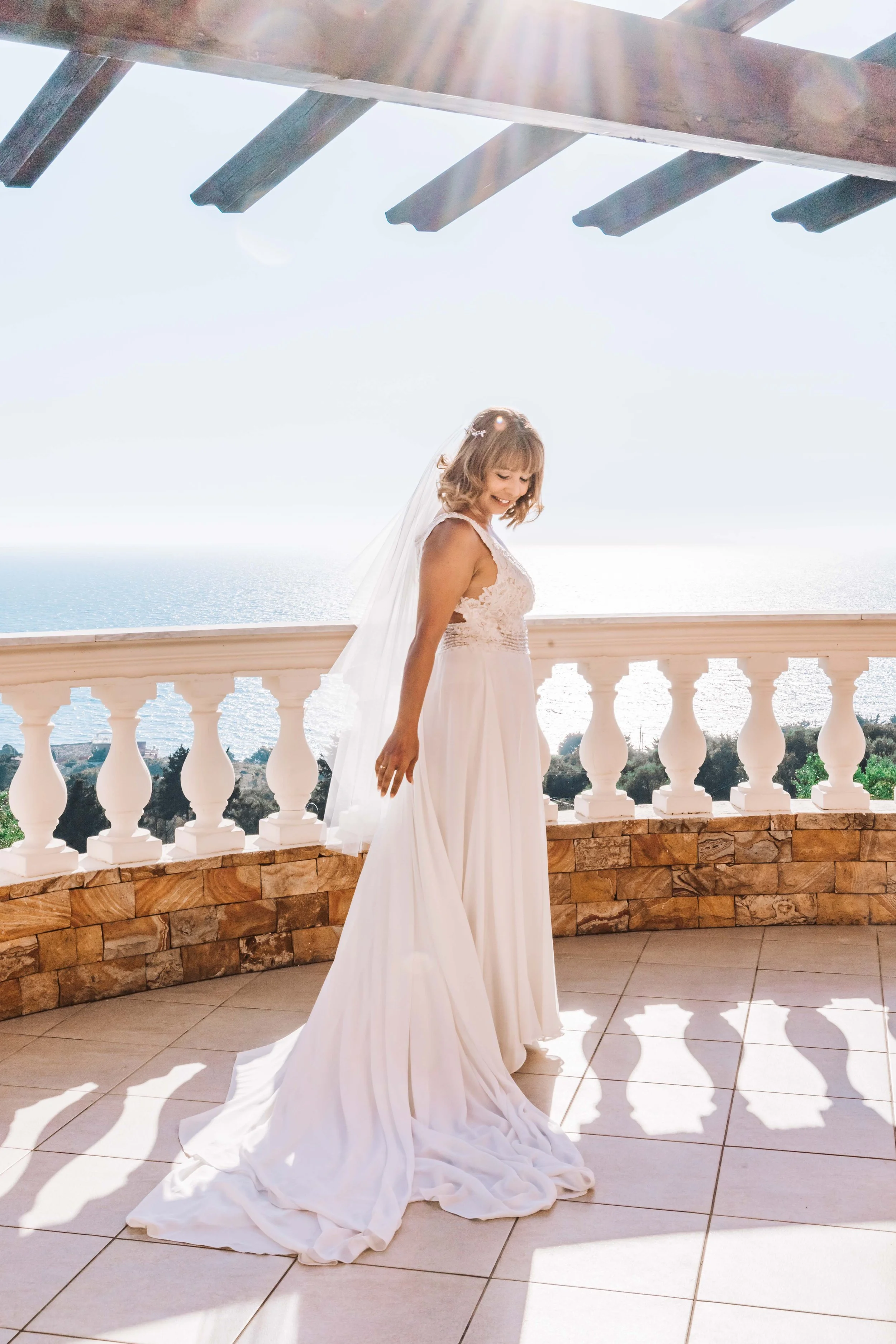 A woman in a white bridal dress with a veil on a balcony with a view of the ocean and blue sky.