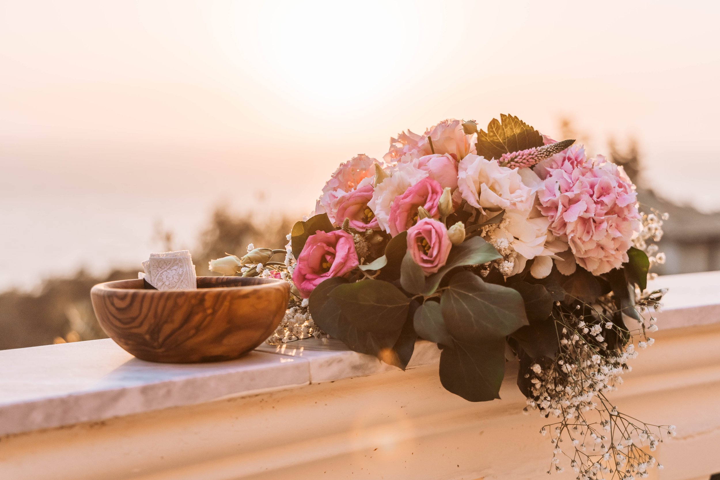 A bouquet of pink and white flowers with greenery resting on a white ledge at sunset, alongside a small wooden bowl with a lace item inside.