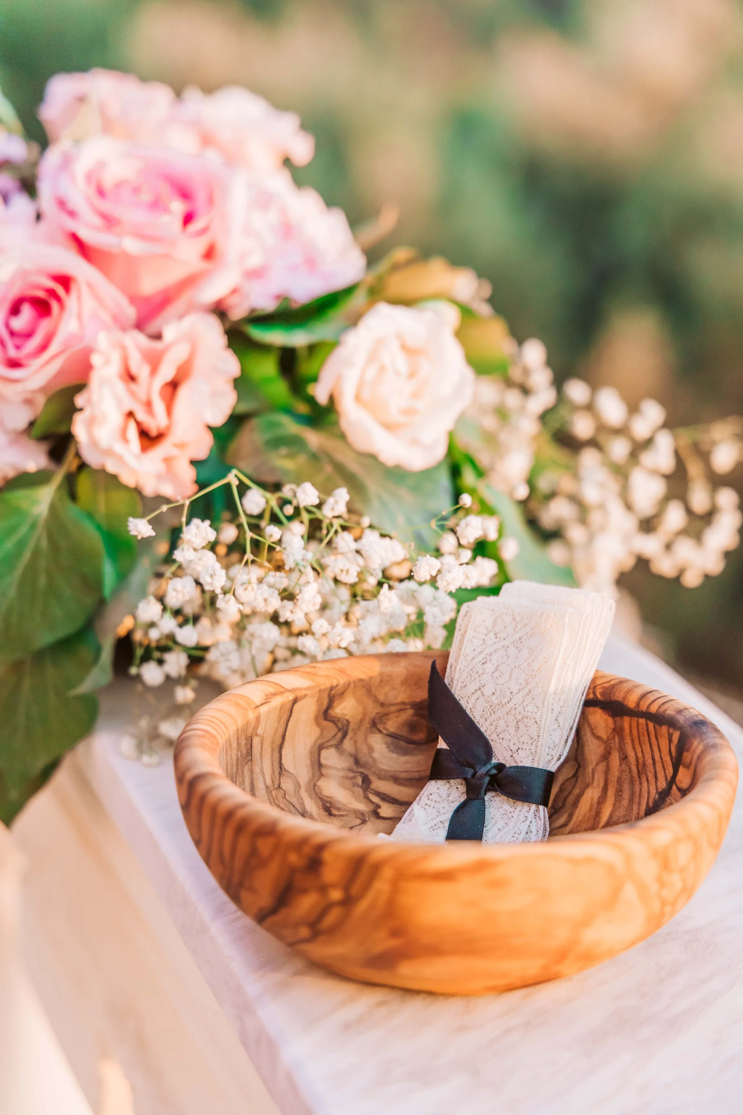 A wooden bowl holding a lace napkin tied with a black ribbon, set on a white surface. Behind it, there are pink and white roses with green leaves and white baby's breath flowers, creating an elegant floral arrangement.