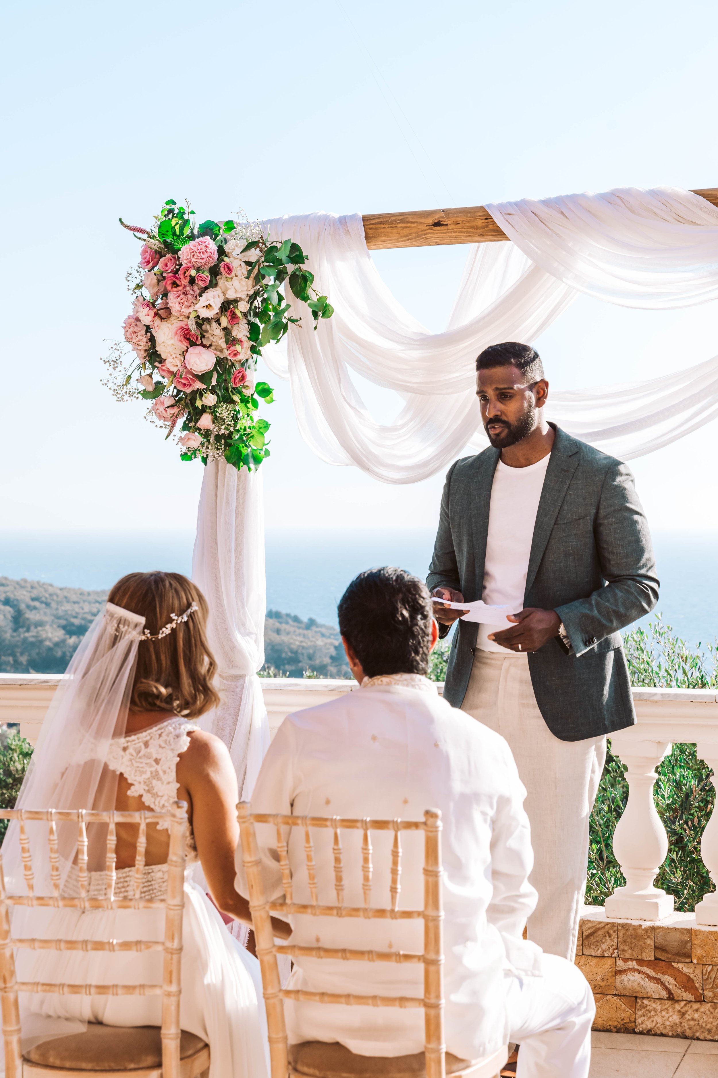 A wedding ceremony takes place outdoors with a man in a gray blazer, a woman in a white dress with a veil, and another man in white sitting before an altar decorated with pink and white flowers and white drapery, overlooking a landscape of trees and 