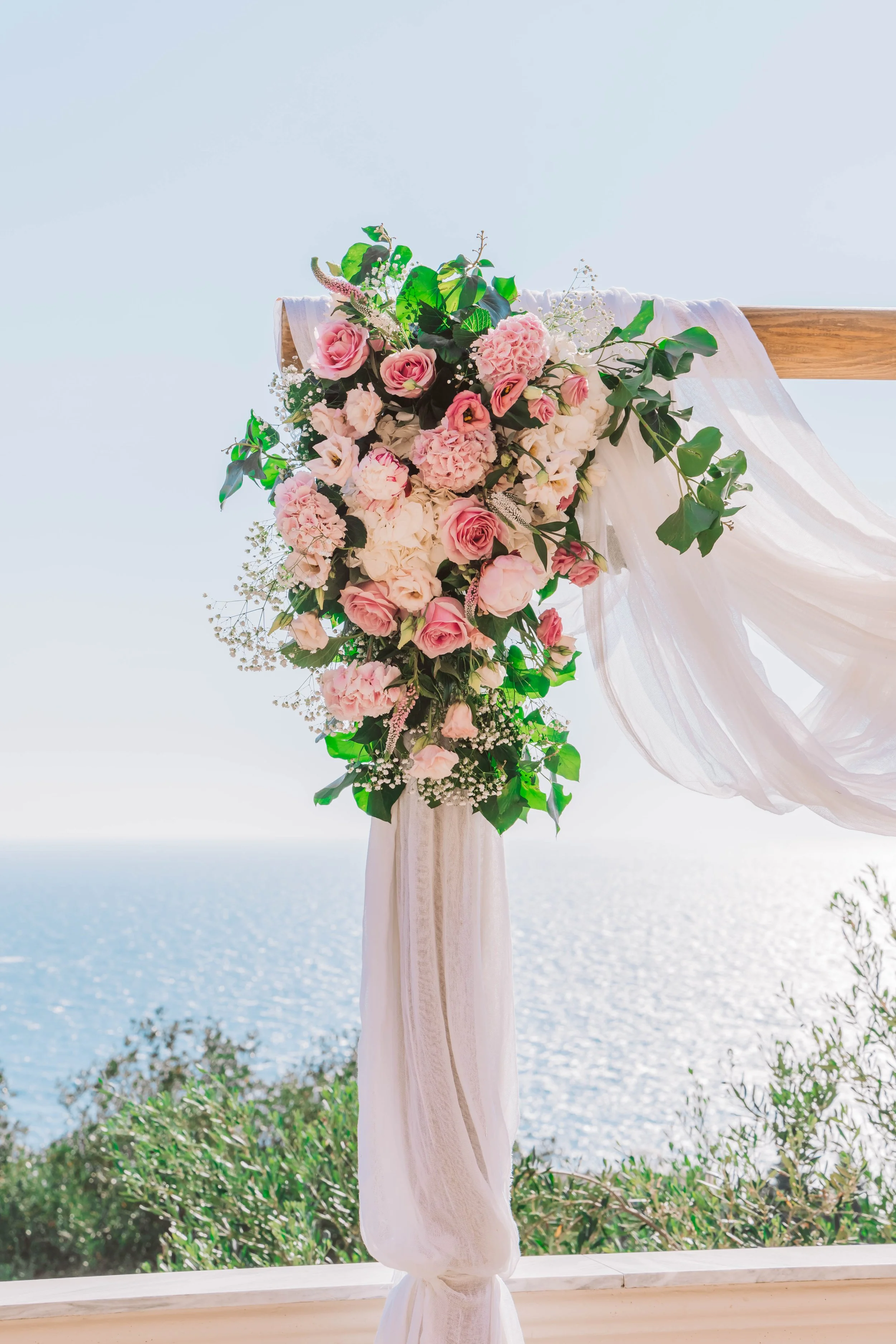 A floral wedding arch with pink and white roses, hydrangeas, and greenery, draped with white fabric, overlooking the ocean under a clear blue sky.