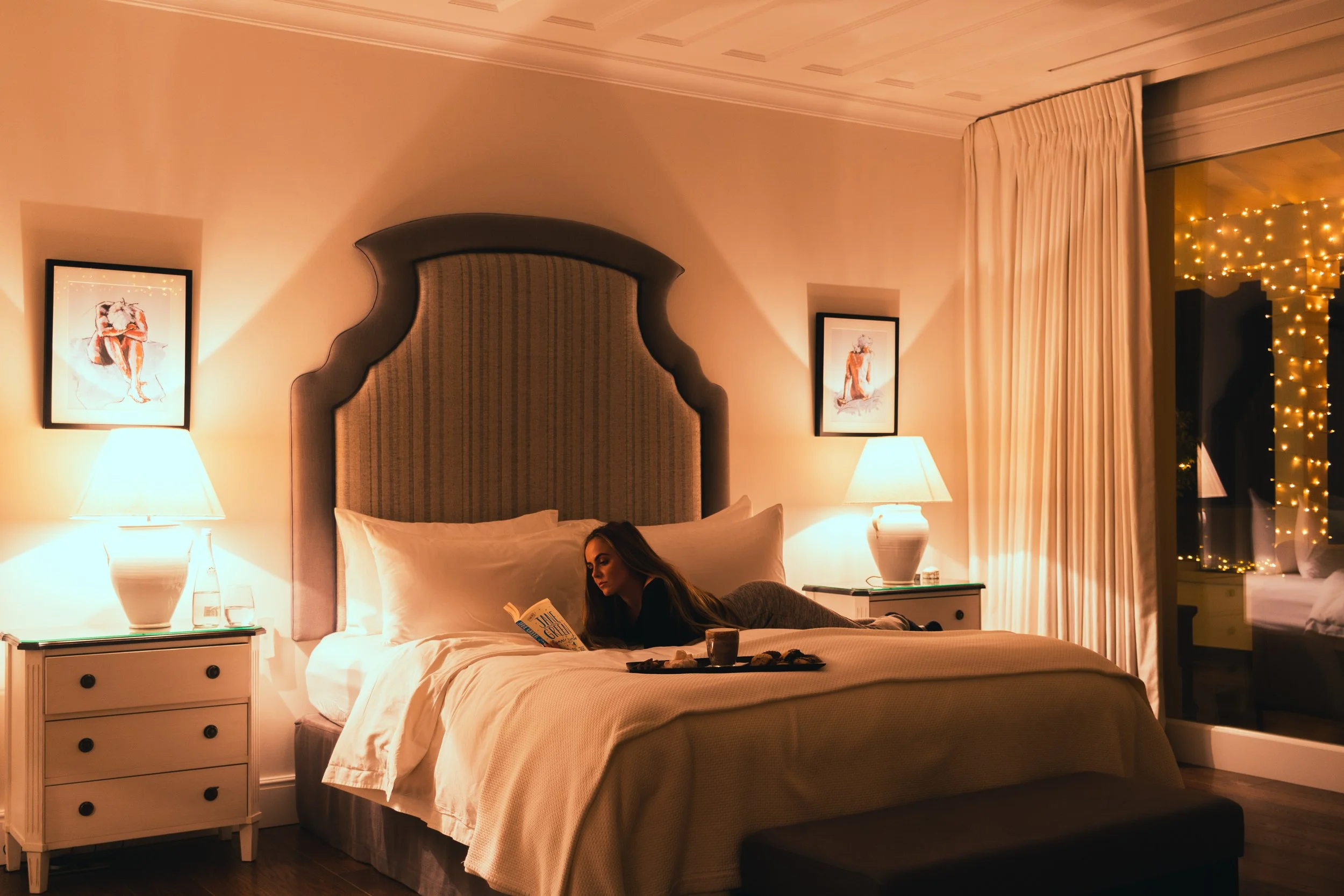 A woman lies on her stomach on a bed in a warmly lit hotel room, reading a book. The room has two nightstands with white lamps, and there are paintings on the wall behind the bed. Large curtains partially cover sliding glass doors leading to a balcon